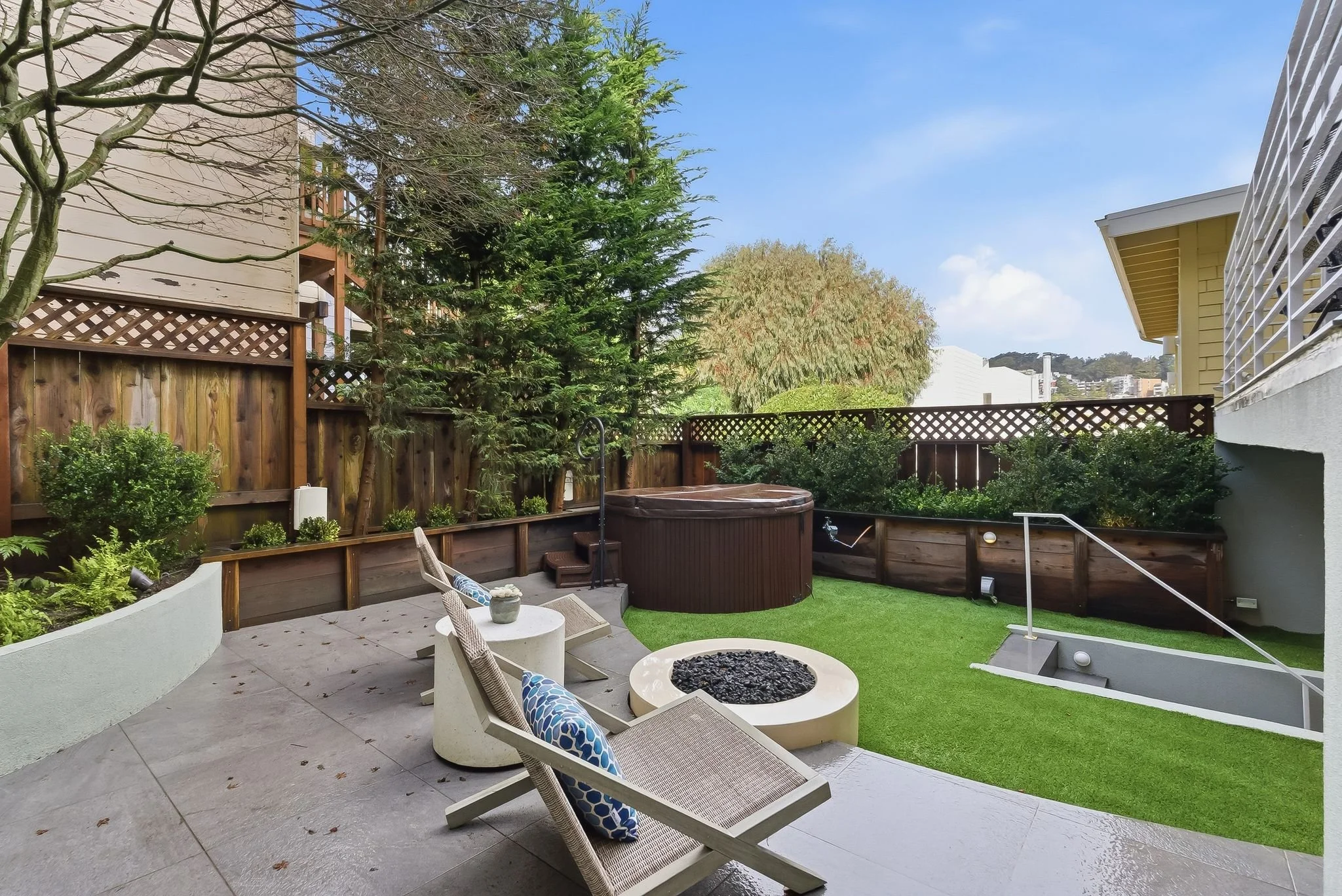 A backyard patio with two chairs, a small round table, a fire pit, a hot tub, and a grassy lawn, enclosed by a wooden fence with trees and shrubs.