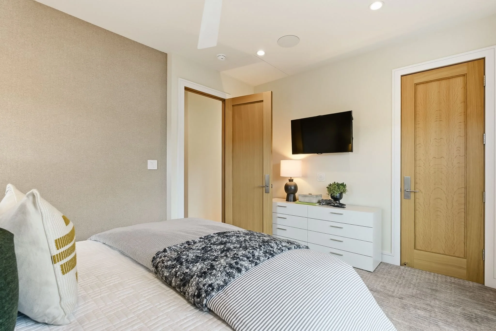 A bedroom with a bed, dresser, television, and two wooden doors, decorated in neutral tones with minimal furnishings.
