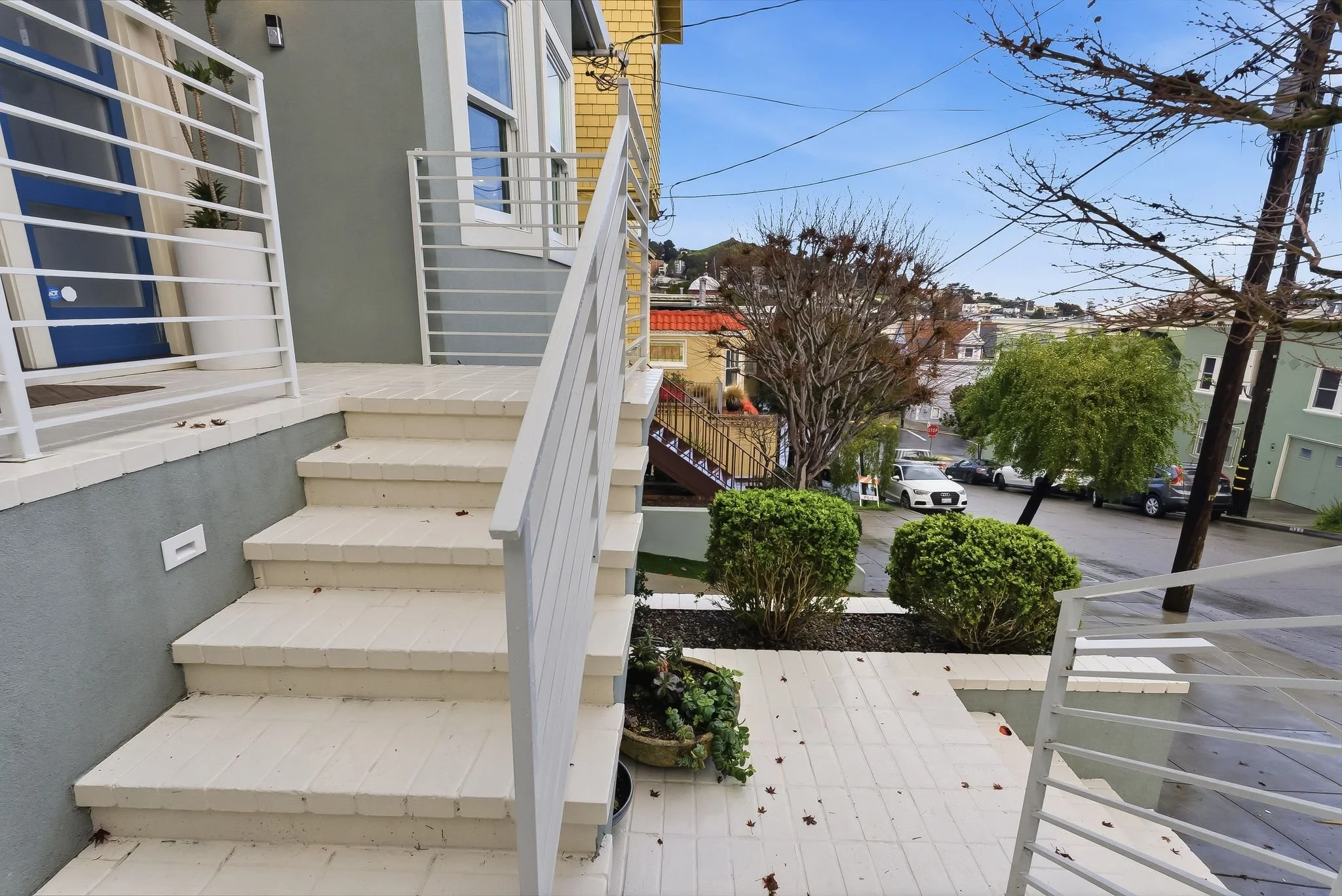 Exterior view of a residential building with stairs and small garden, showing trees, parked cars, and neighboring houses on a partly cloudy day.
