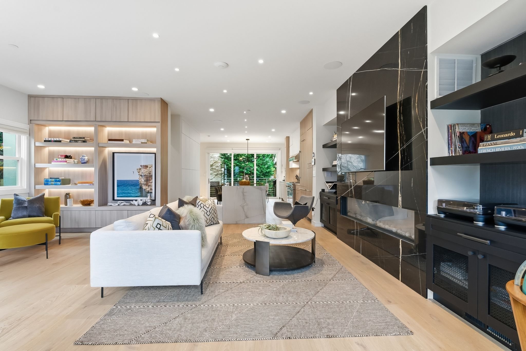 Modern living room with a white sectional sofa, round coffee table, and built-in shelves with a television and decor. Open kitchen and dining area in the background, with large windows and natural light.