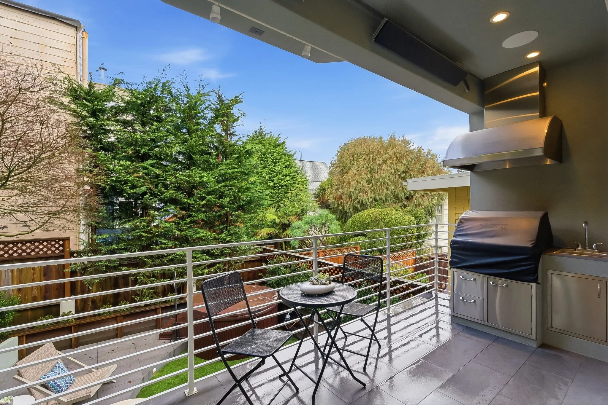View of a balcony outdoor space with a small round table and two chairs, a built-in grill, and lush green trees and foliage in the background.