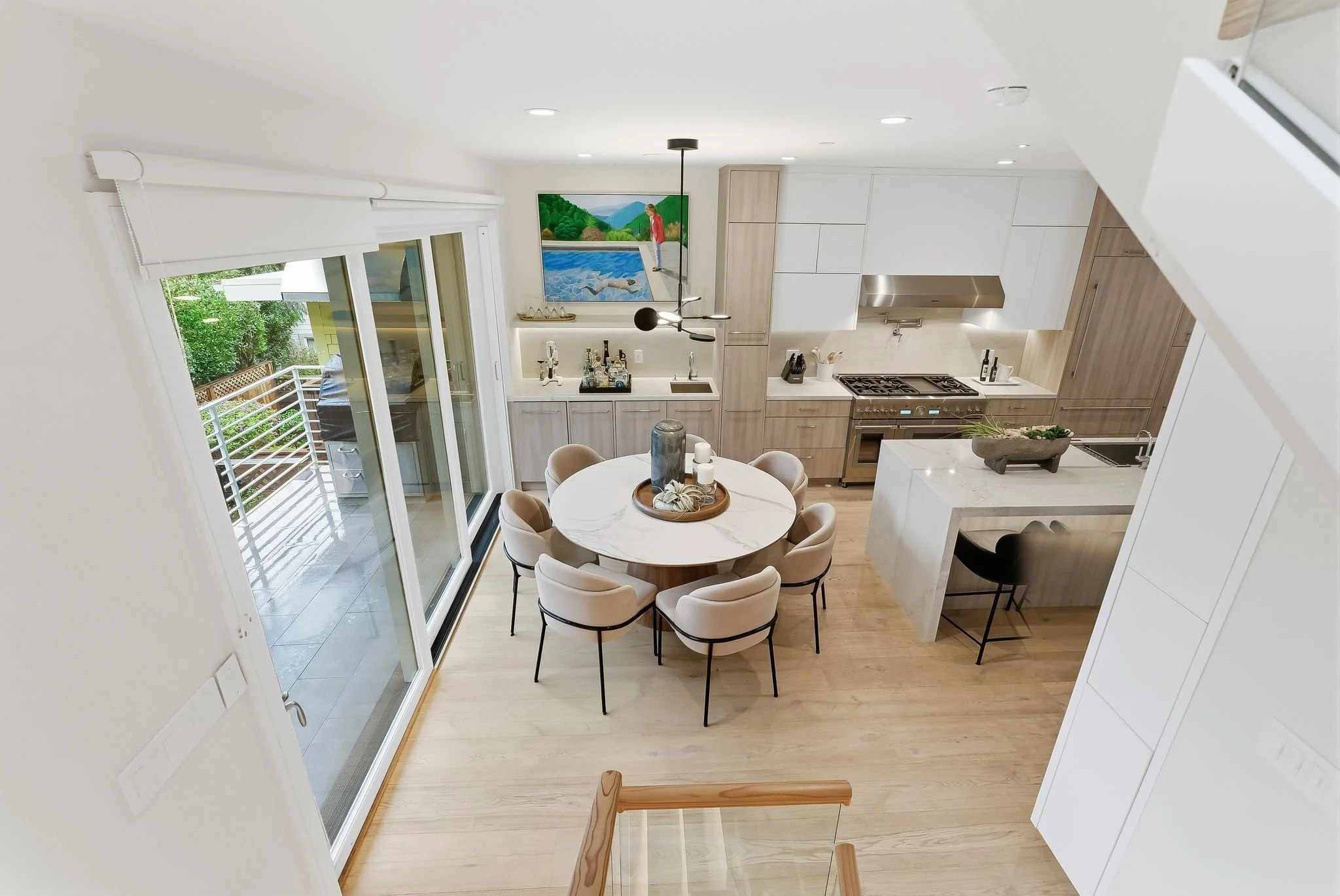 Modern kitchen with white cabinetry, a round dining table with beige chairs, a sliding glass door leading to a balcony, a wall-mounted painting of a person beside a river, and light wood flooring.