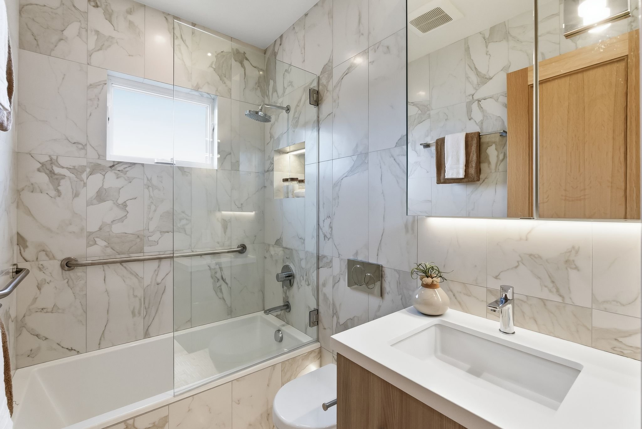 Modern bathroom with marble walls, a small window, a shower with glass door, a white sink with a wooden cabinet, a plant in a vase, and a large mirror with built-in lighting.