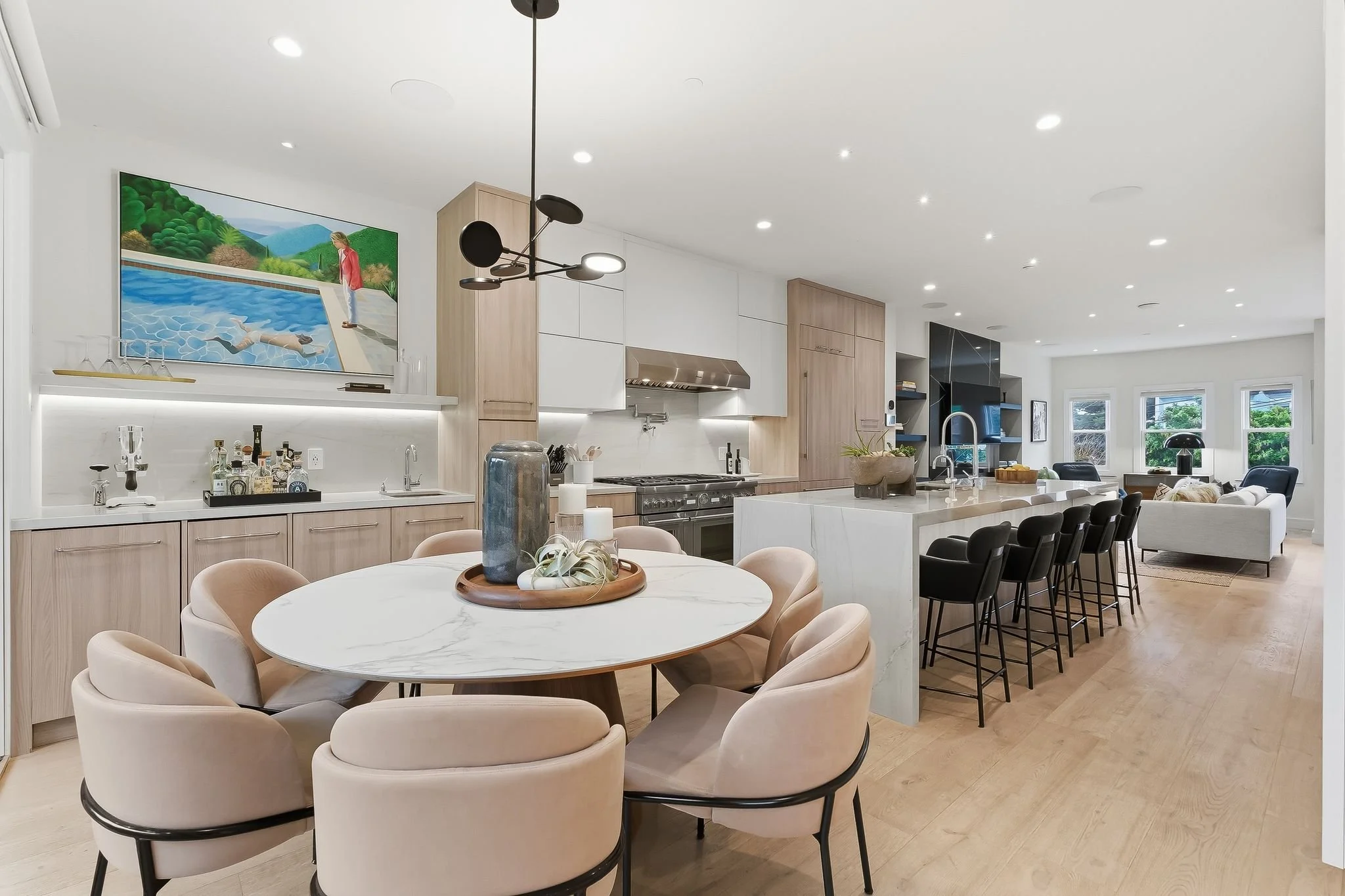 Open-concept kitchen and dining area with light wood cabinets, white countertops, and a marble dining table with beige chairs. A large modern chandelier hangs above the dining table. The kitchen features black barstools at the island, a stainless ste