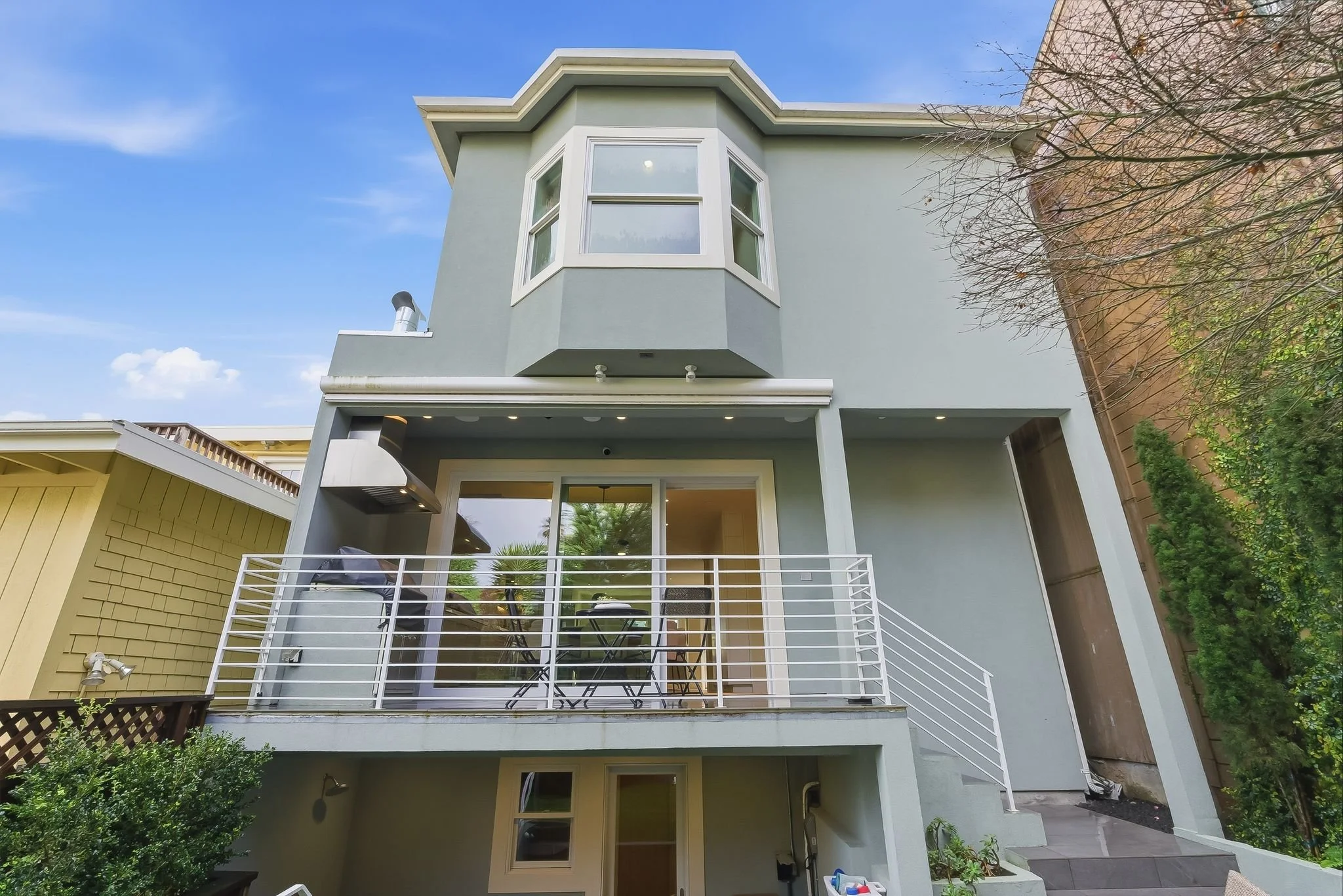 A modern multi-story house with a light green exterior, a balcony with white railing, and large windows under a blue sky.