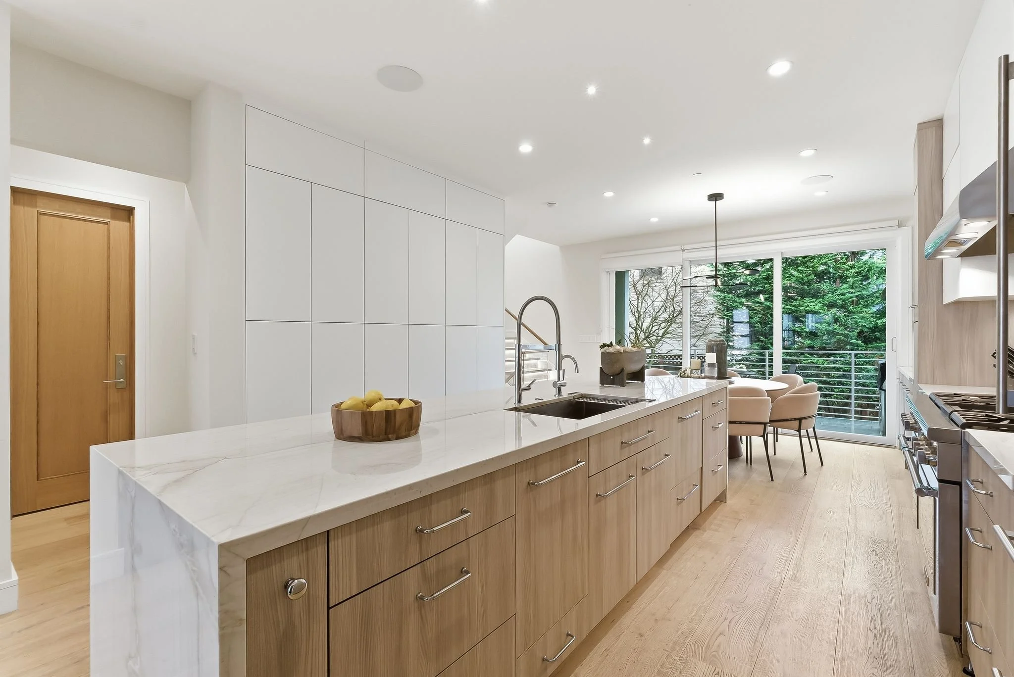 Modern kitchen with light wood cabinetry, white marble countertops, and a dining area with chairs near large glass doors opening to a balcony with trees outside.