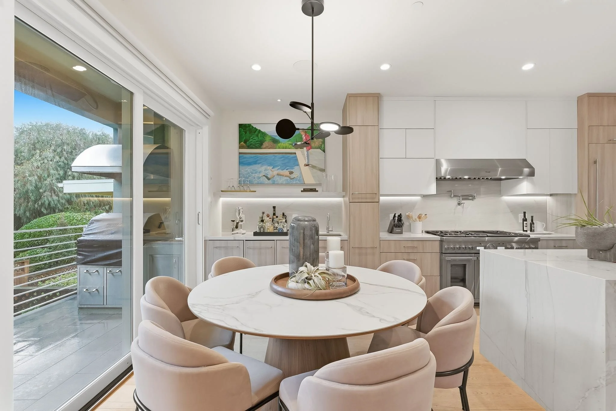 Modern kitchen with a round marble dining table, beige chairs, white cabinetry, stainless steel appliances, large sliding glass door leading to an outdoor area, and decorative candles and plants on the table.