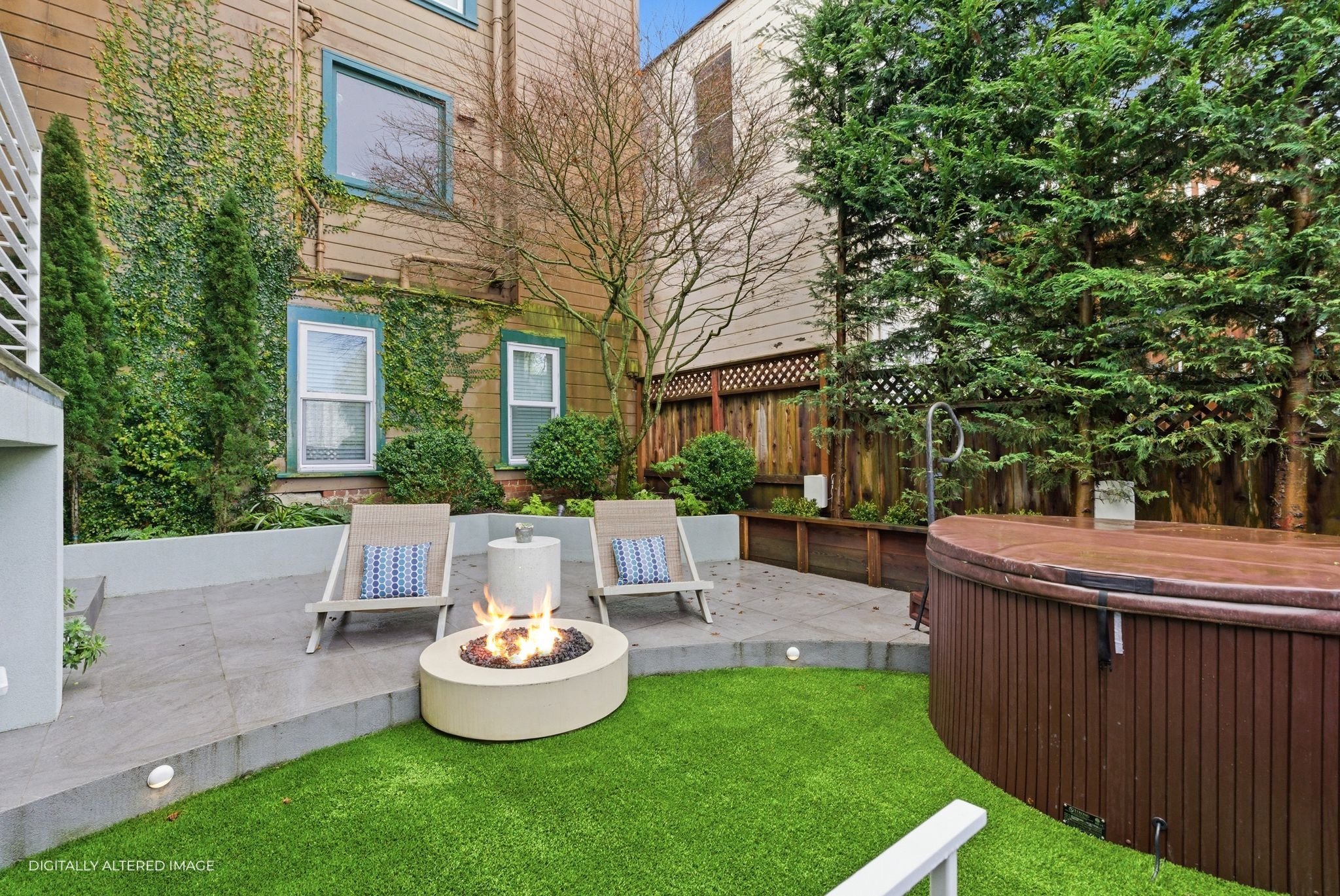 Urban backyard featuring a fire pit with two chairs, a hot tub, and surrounding greenery including trees and shrubs.