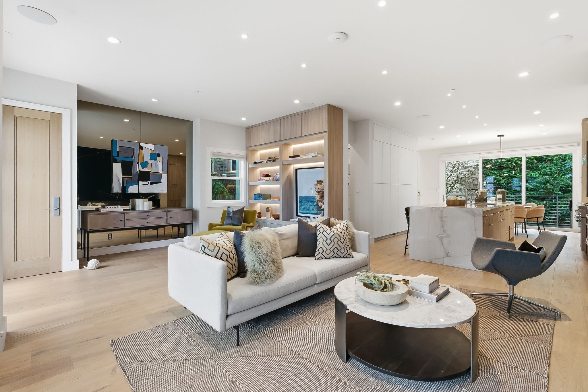 Modern open-concept living room with white sofa, decorative pillows, a gray swivel chair, and a round marble coffee table, adjacent to a kitchen with a large window, light wood flooring, and built-in shelving.