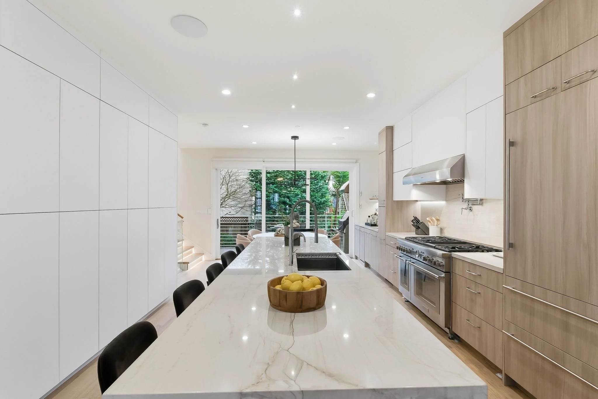 Modern kitchen with white and light wood cabinetry, a large marble island with black chairs, and stainless steel appliances, including a stove. A bowl of lemons is on the island, and there is a view of a balcony with trees outside.