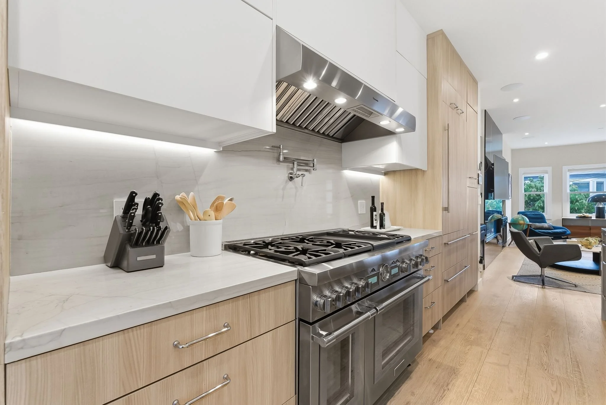 Modern kitchen with white and wooden cabinets, marble countertop, stainless steel stove, and kitchen utensils on the counter, with a glimpse of a living room in the background.
