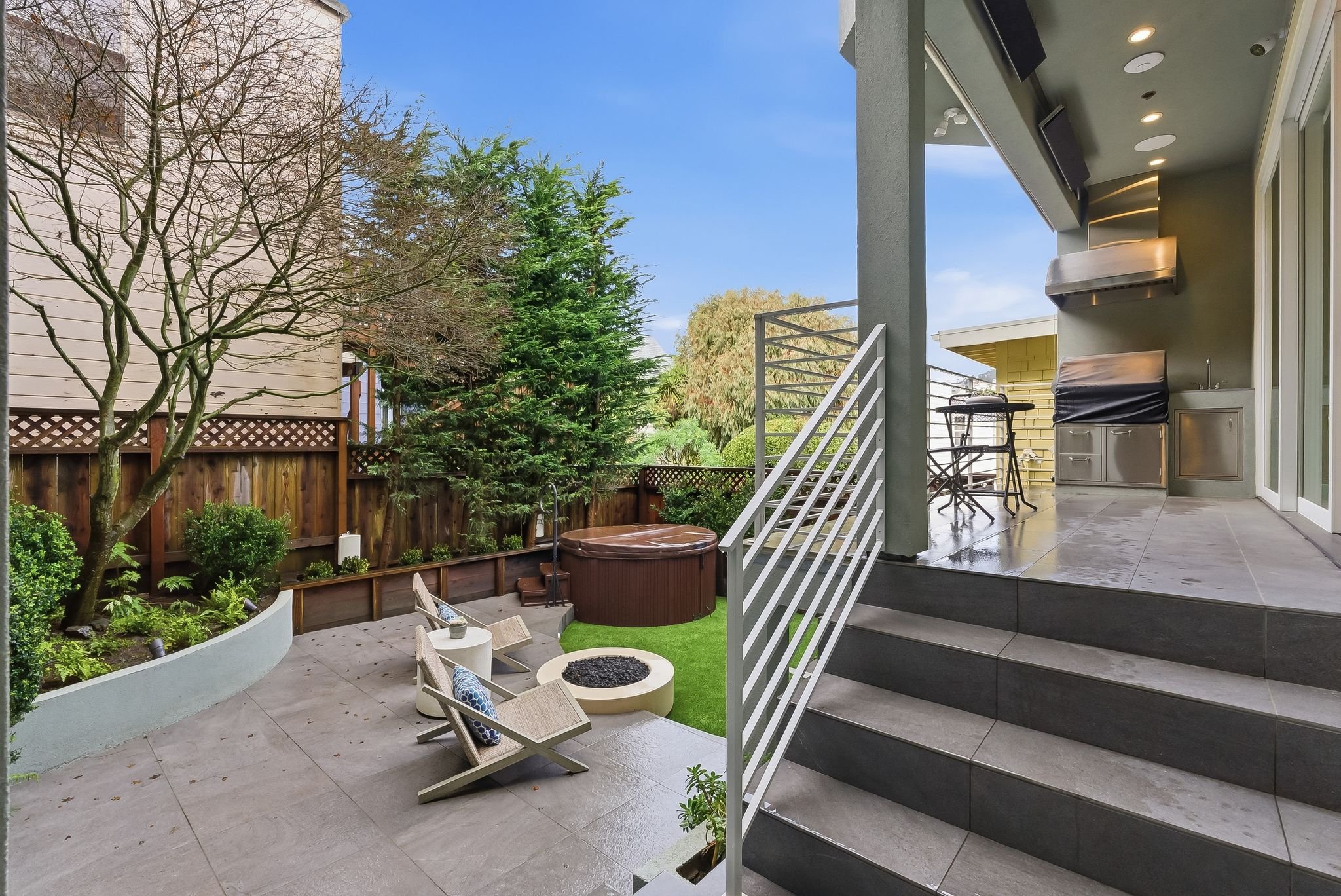 View of a backyard patio area with a hot tub, seating, a fire pit, and a barbecue, enclosed by a wooden fence and surrounded by trees and greenery.
