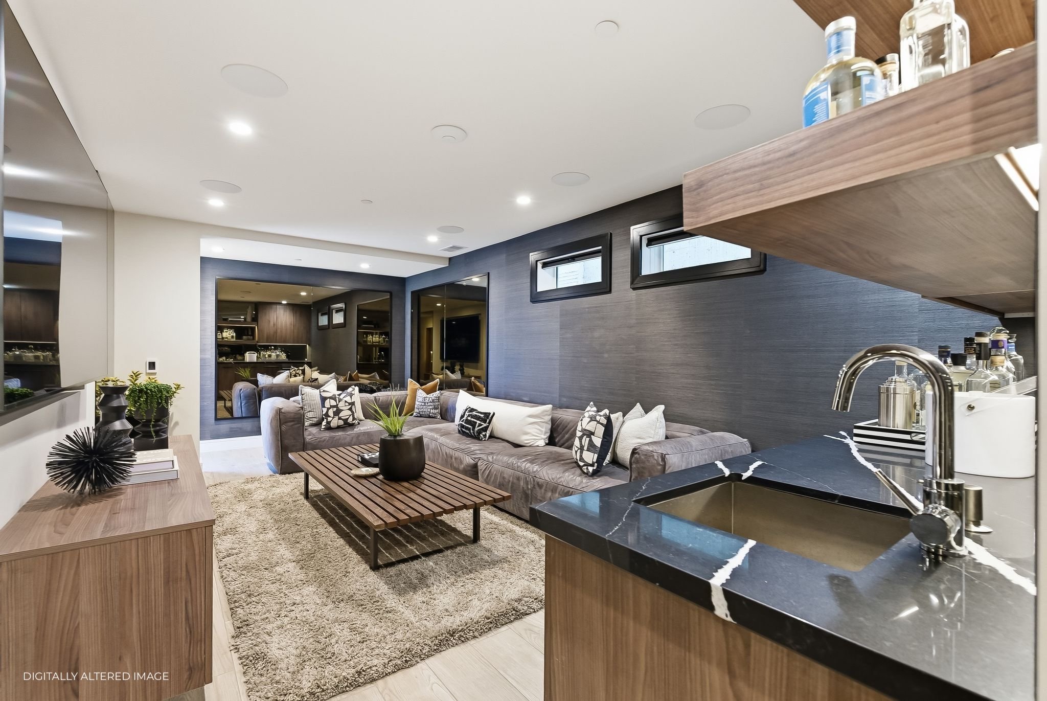 Modern living room with a large sectional sofa, decorative pillows, a coffee table, and a plush area rug. There are small windows on the dark accent wall and a kitchenette with bottles and a sink in the corner.