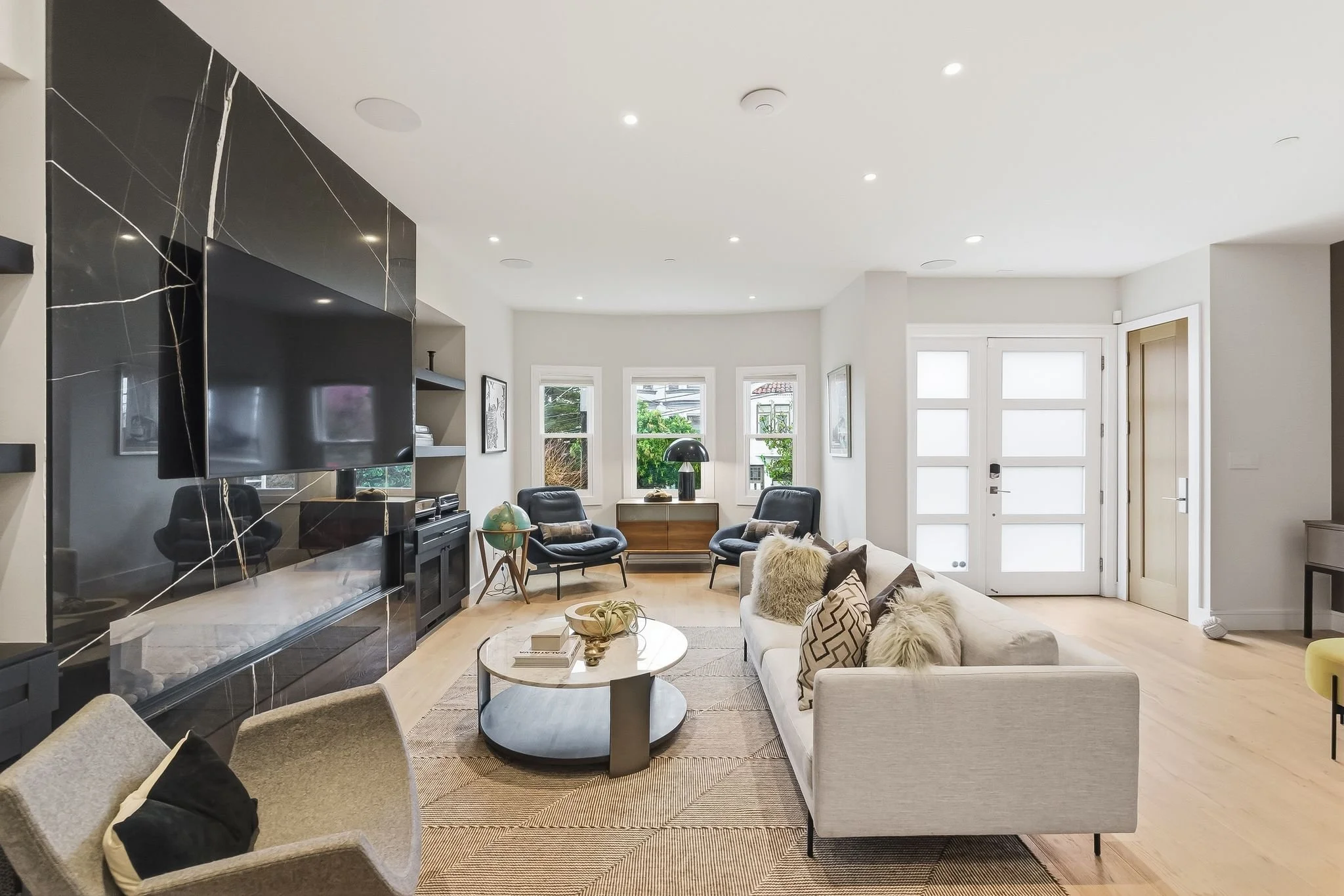 Modern living room with a large flat-screen TV on a black marble wall, a beige sofa with pillows, two black armchairs in front of a bay window, a round coffee table with decorative items, and a globe on a small side table. Bright natural light fills 