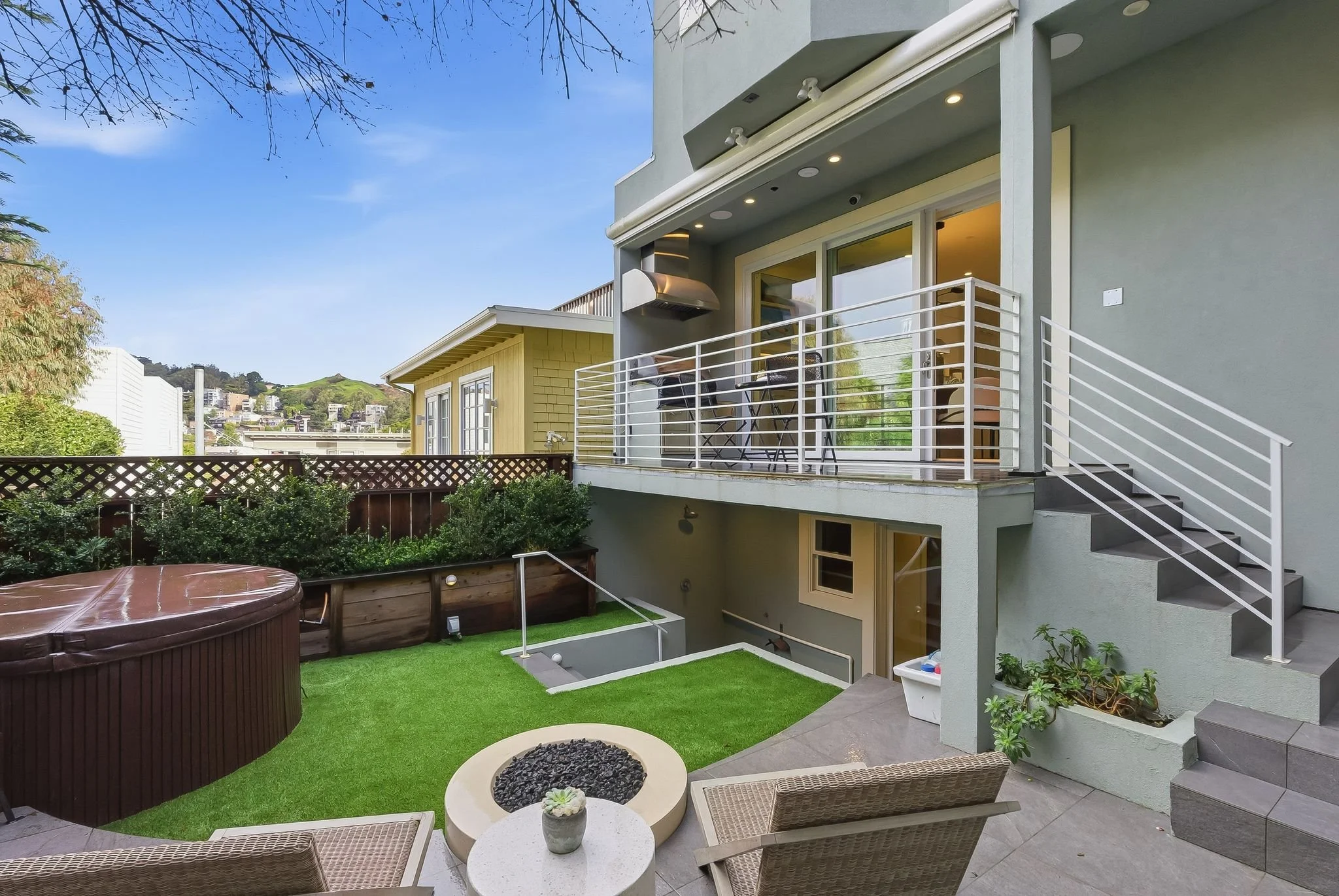 Outdoor patio with wicker chairs, small round table with a potted plant, fire pit with rocks, hot tub, elevated balcony with glass doors, plants, and stairs, green lawn, and a garden area with bushes, fence, and neighboring houses in background.