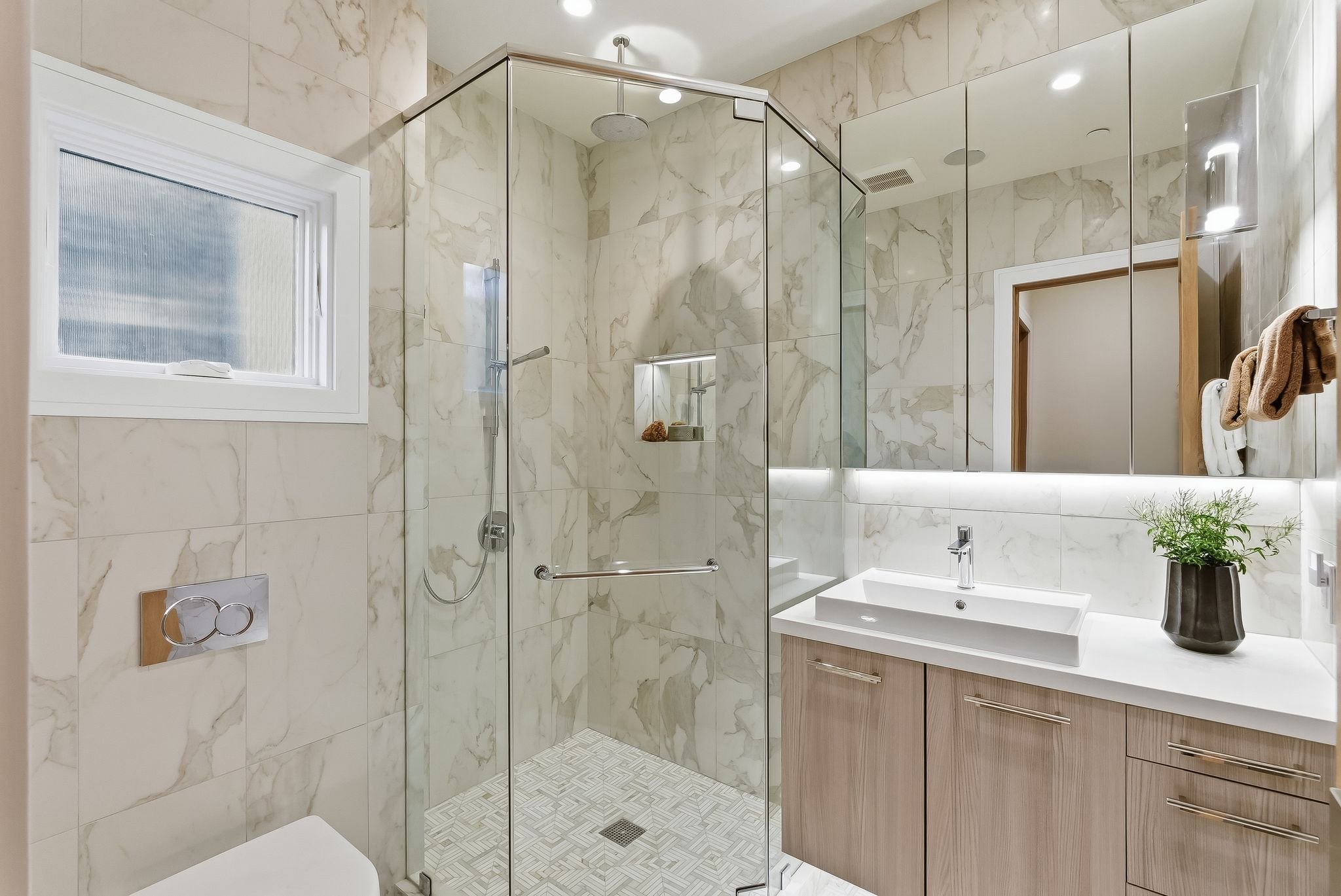 Modern bathroom with marble tiles, glass shower enclosure, white vanity with wooden cabinet, large mirror with backlighting, potted plant, window, towel rack, and ceiling lights.