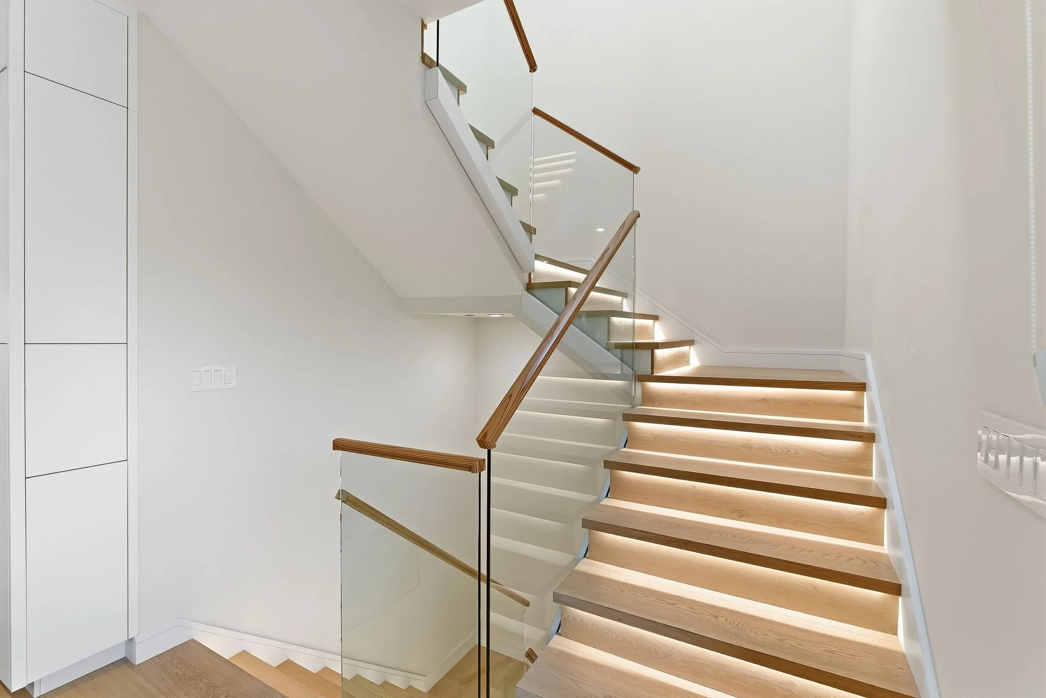 Modern staircase with wooden steps, glass railing, and white walls.