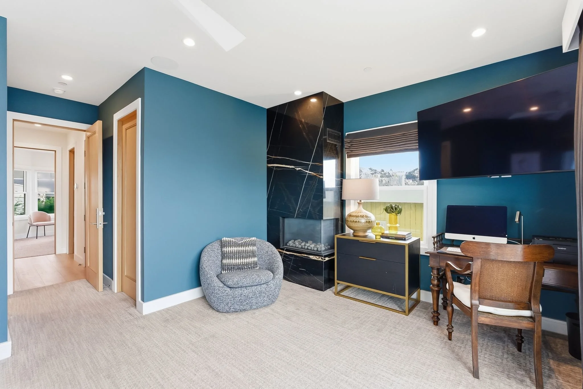 Living room with blue walls, a black marble fireplace, a gray rounded chair with a cushion, a black and gold side table with a lamp and decorative items, a window with blinds, a large flat-screen TV, an iMac on a wooden desk, and a chair in the corne