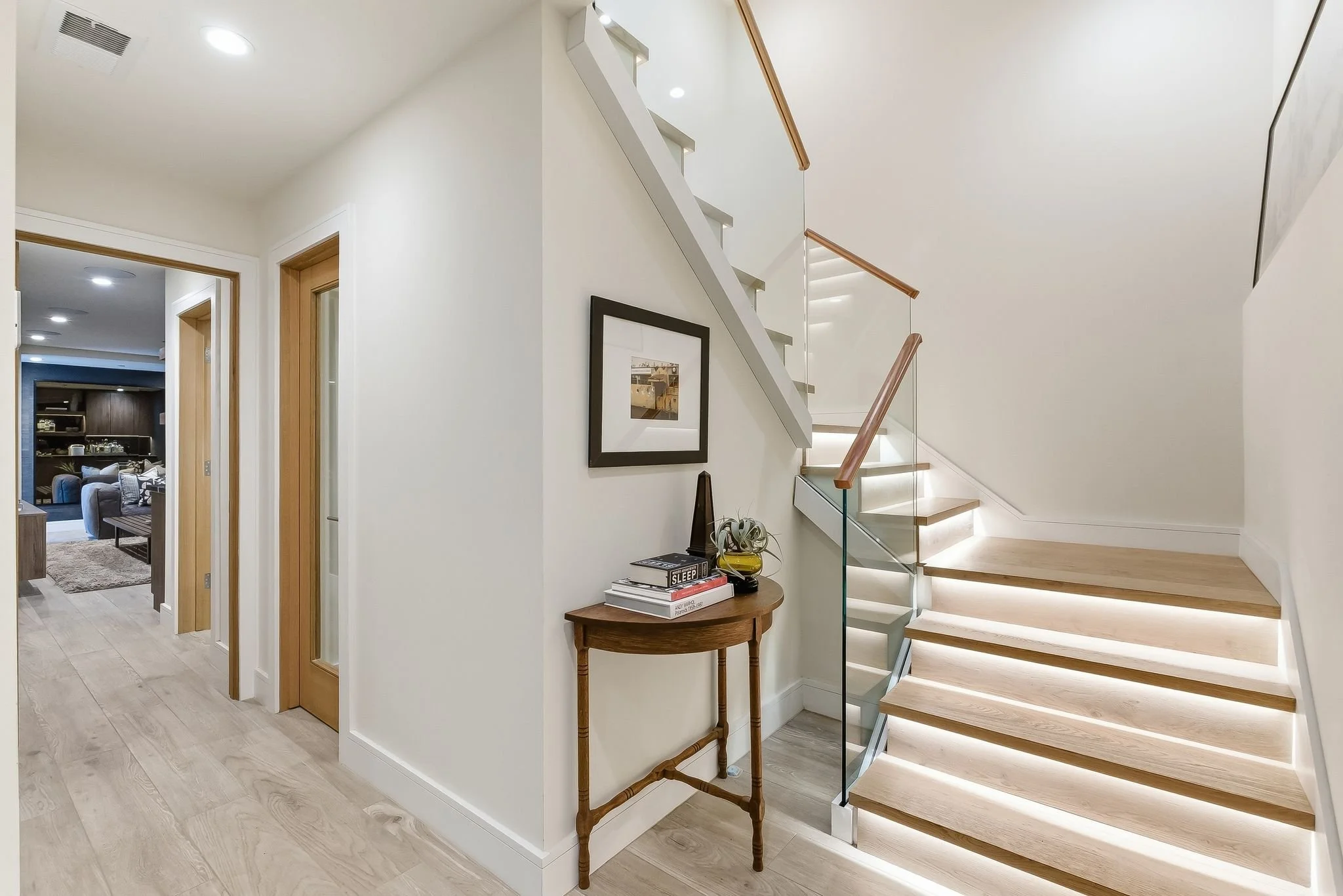 Interior of a modern home with a staircase featuring glass railing and wooden steps, a small wooden table with books and a plant, and a hallway leading to a living room.