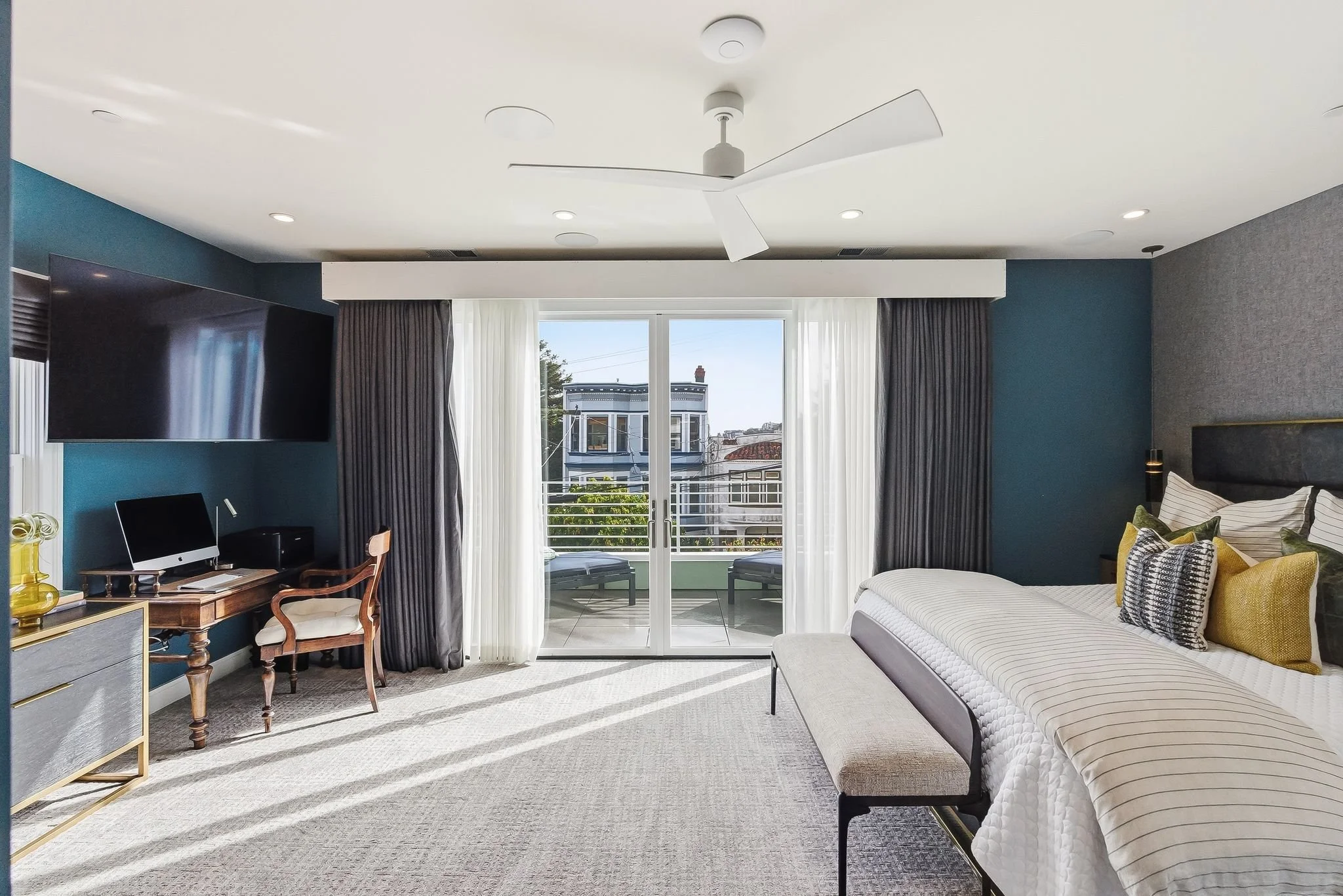 Modern bedroom with large sliding glass doors leading to balcony, a bed with multiple pillows, a wooden desk with a TV and computer, and a ceiling fan.