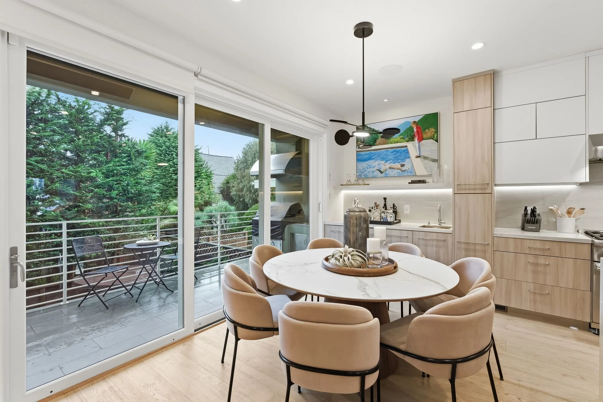 Modern kitchen and dining area with a sliding glass door leading to an outdoor balcony with two chairs and a small table, overlooking greenery.