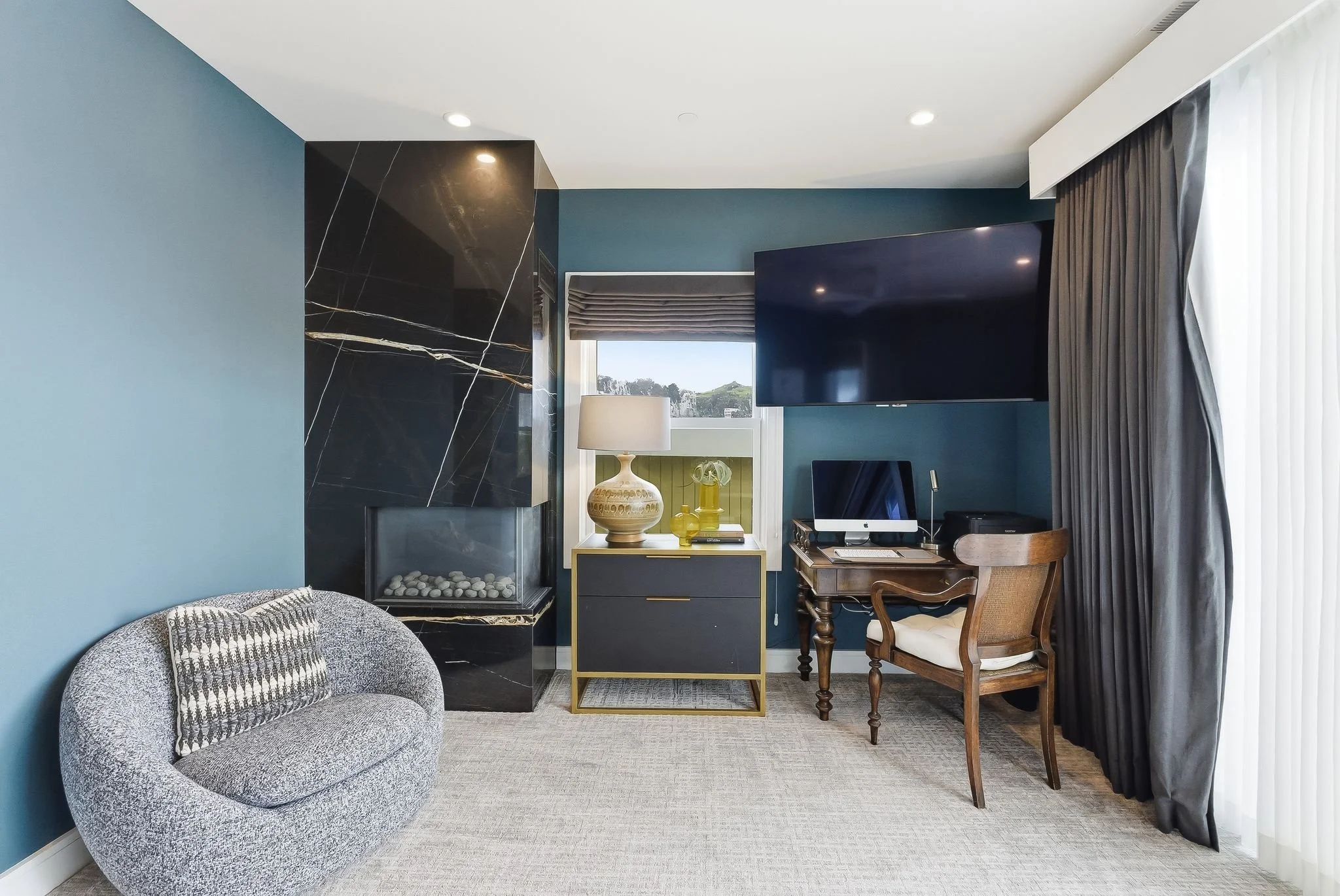 Cozy living room with a gray armchair, a black marble fireplace, a window with a beige shade, a dark sideboard with a yellow vase, a lamp, and decorative glass containers, a large TV mounted on a blue wall, a wooden desk with a computer, and gray cur