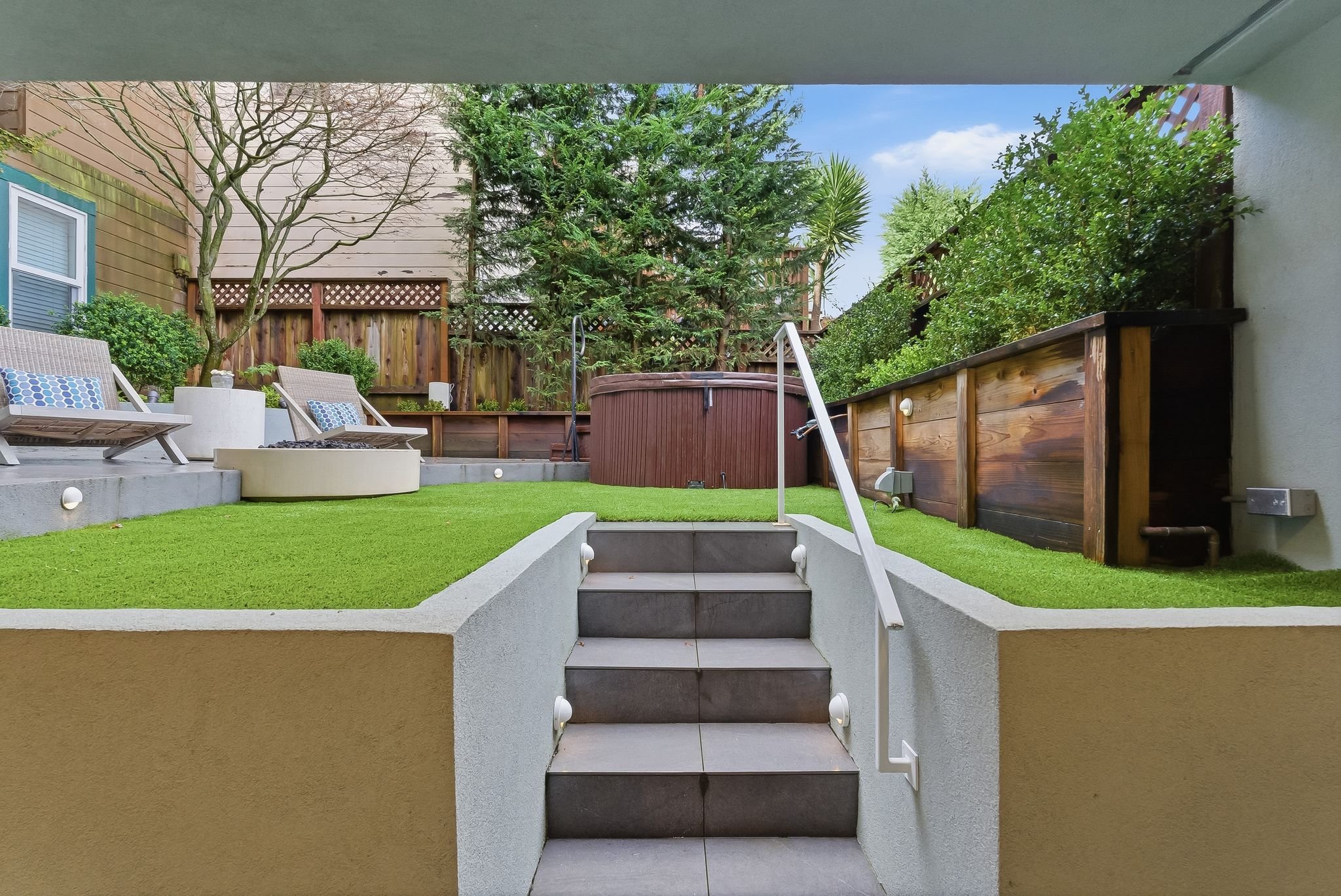 View of a backyard patio with artificial grass, celling stairs, wooden fence, trees, bushes, a hot tub, and outdoor furniture.