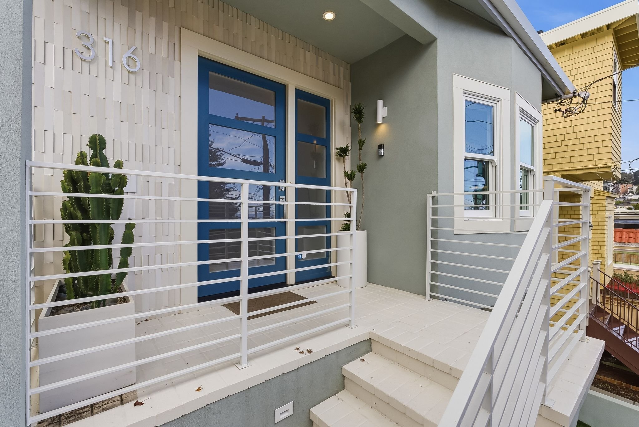 Front porch of a modern house with gray and yellow walls, white metal railing, potted cactus, and a blue door, during daytime.