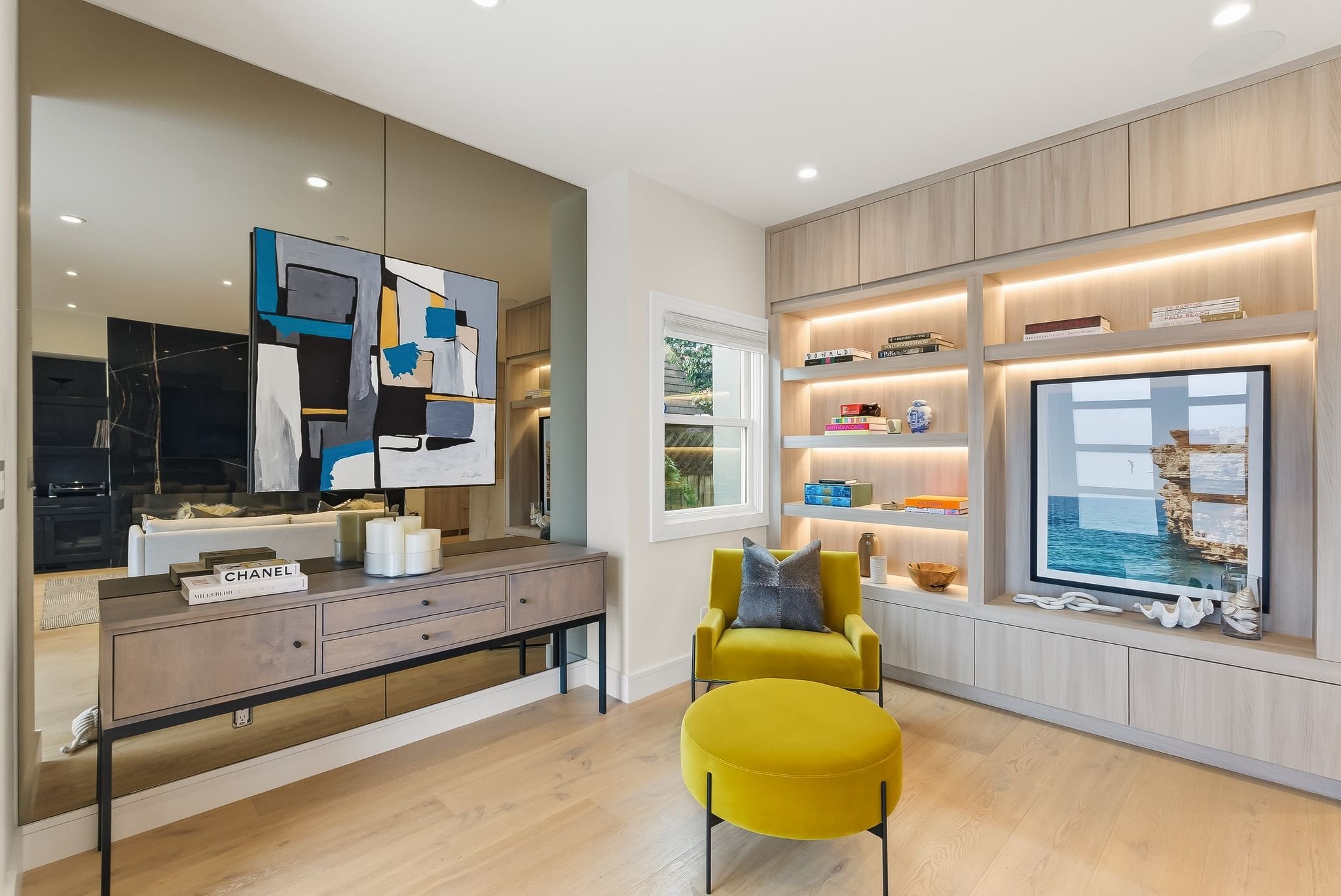 Living room with yellow armchair and round ottoman, built-in wooden bookshelf, large window, abstract artwork on mirror wall, console table with books and candles, TV with seaside view reflection.