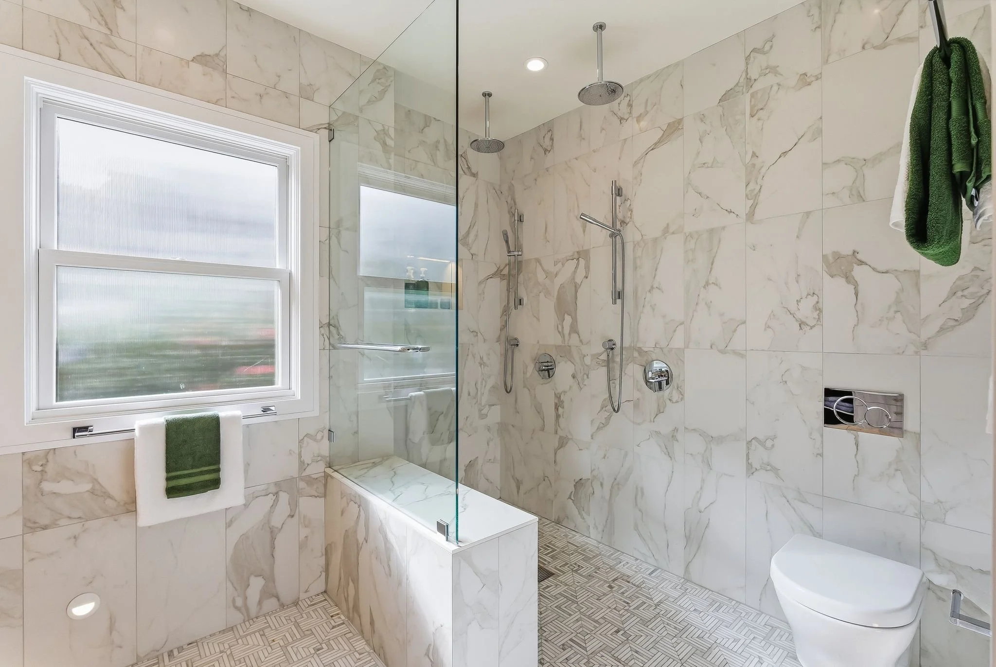 Modern bathroom with marble walls, window with white trim, glass shower with dual showerheads, green towel hanging on wall, toilet, and beige patterned floor.