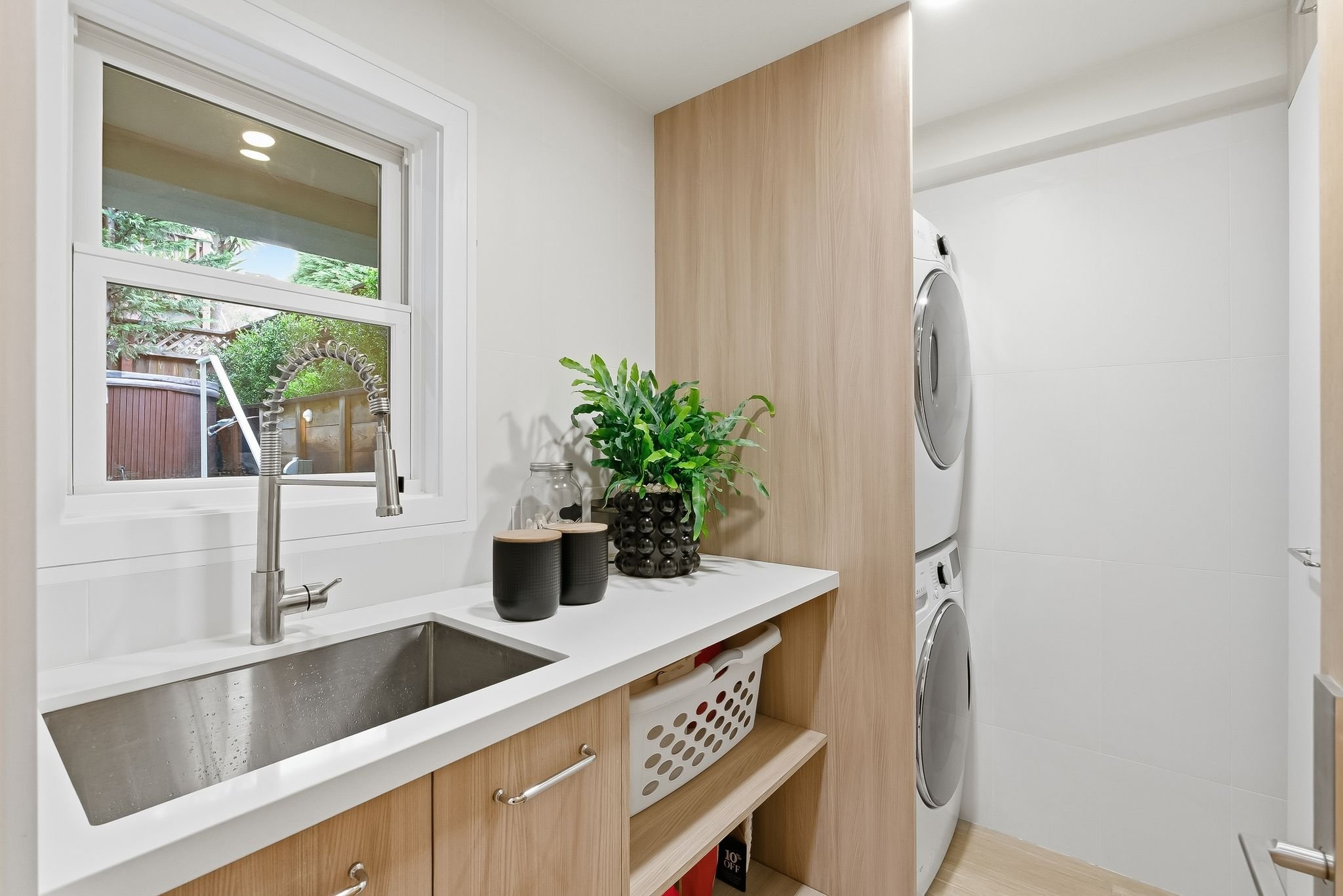 Laundry room with a sink, window, potted plant, black containers, and a stacked washer and dryer behind a wooden partition.