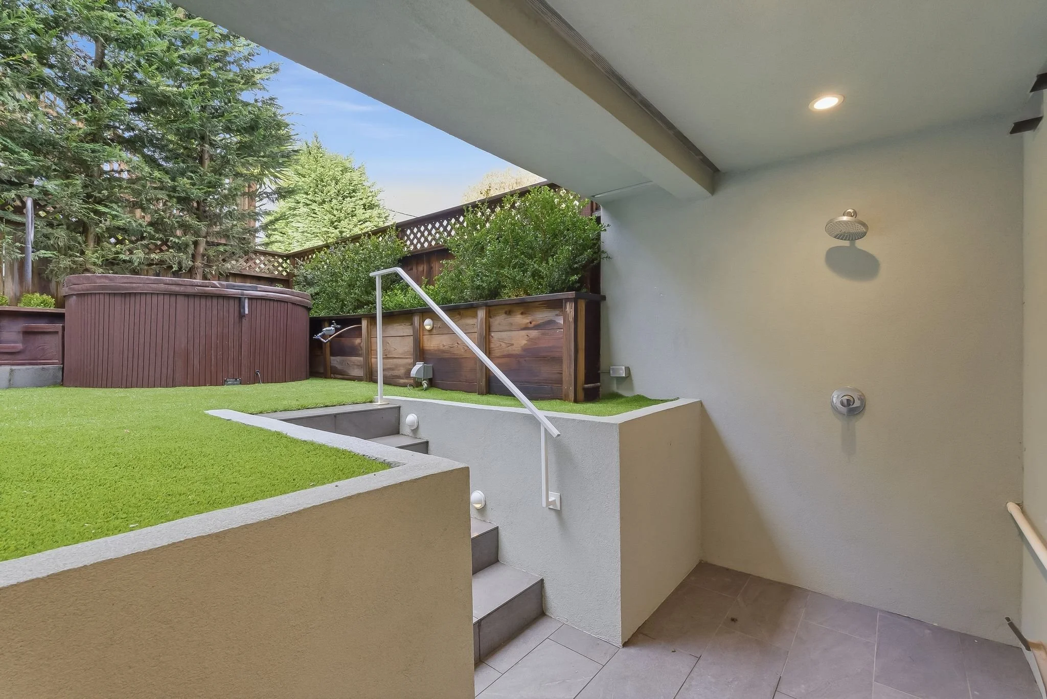 Outdoor patio with steps leading to a grassy area, a Jacuzzi hot tub, and a wooden privacy fence, with trees in the background.
