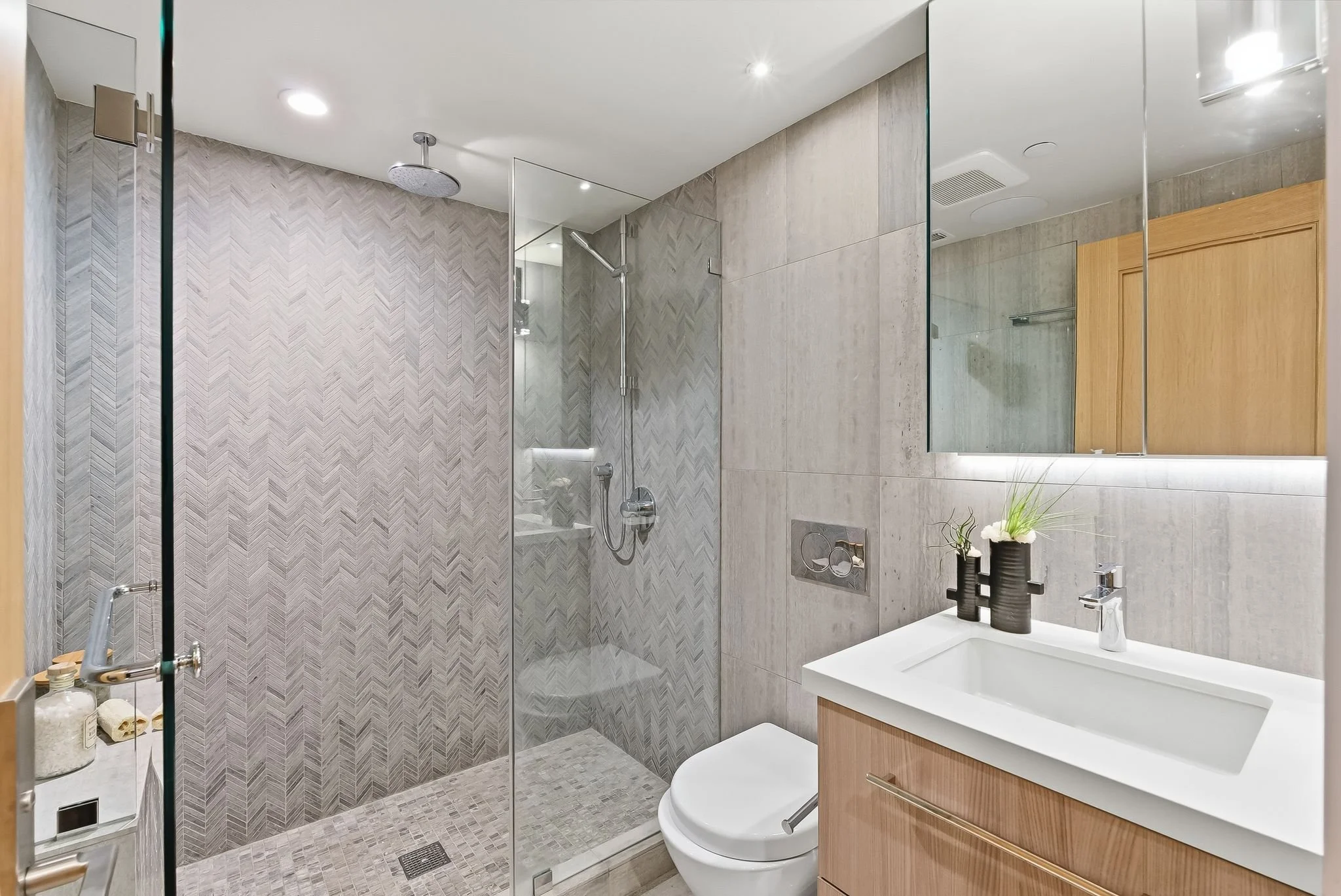 Modern bathroom with glass-enclosed shower, wall-mounted toilet, and white vanity with sink, decorated with black vases and green plants.