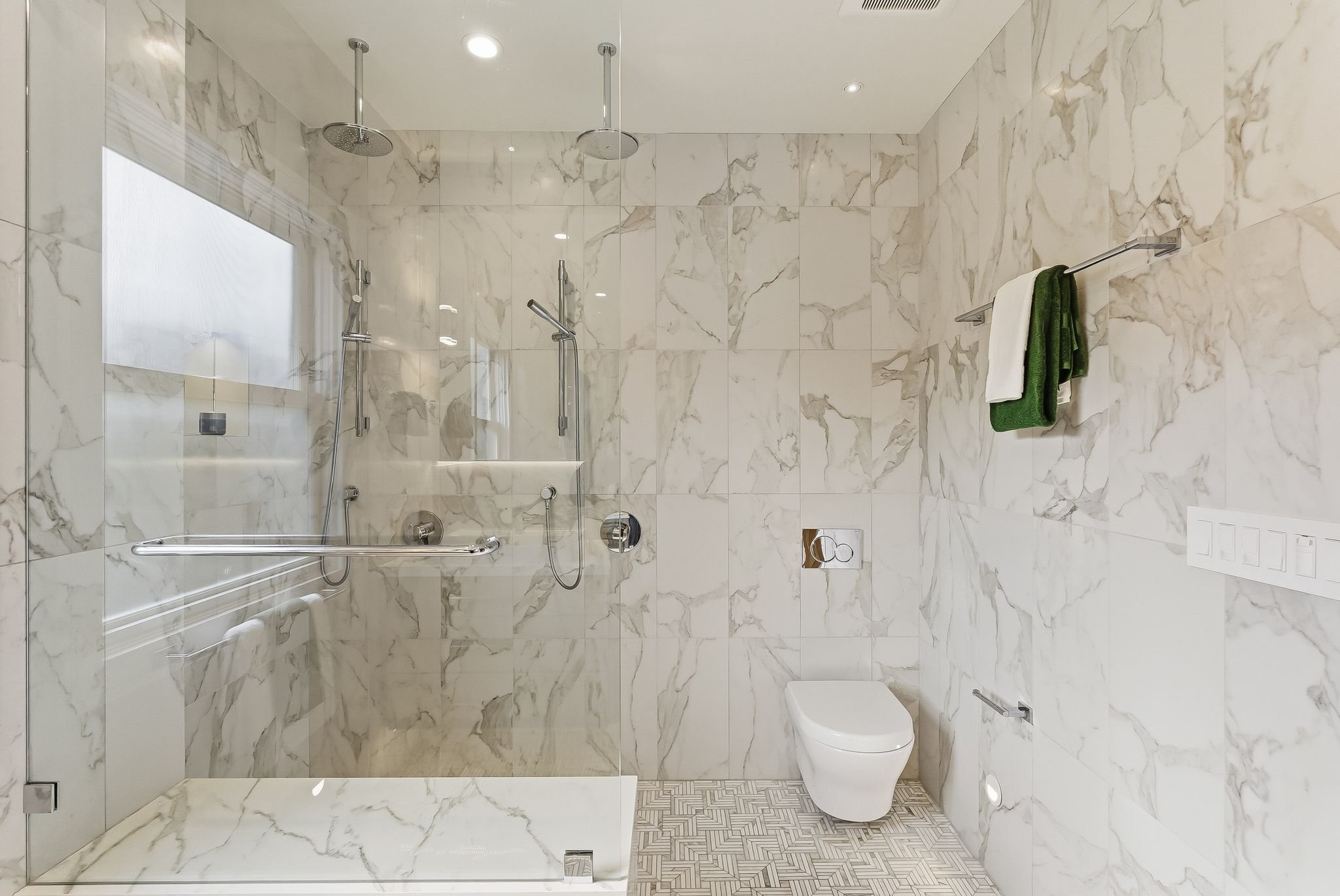 Modern bathroom with marble walls, a glass-enclosed shower featuring multiple shower heads, a wall-mounted toilet, and a towel rack with green and white towels.