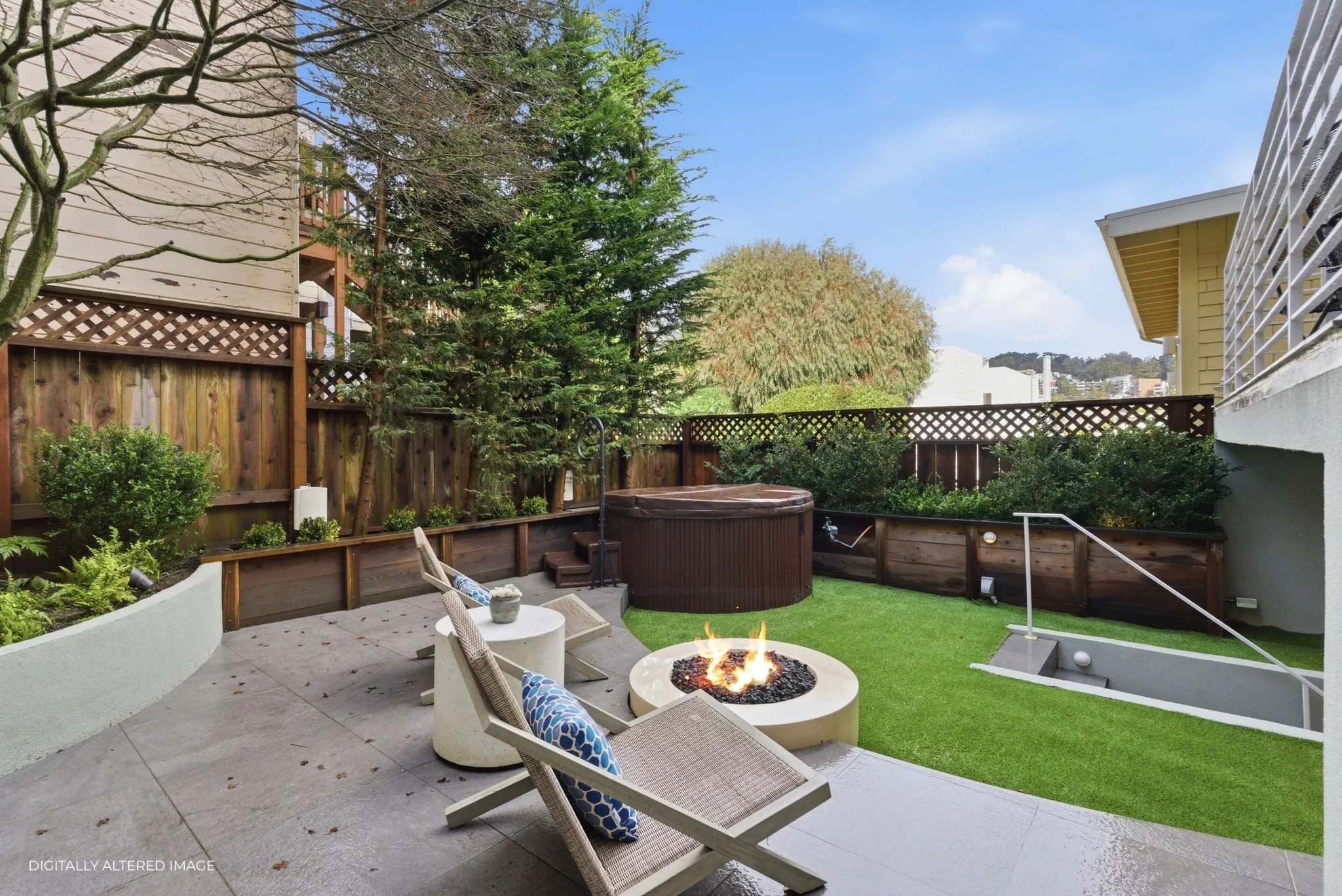 A backyard patio with a fire pit, outdoor chairs with pillows, a hot tub, and a grassy area enclosed by a wooden fence, with trees and neighboring houses in the background.