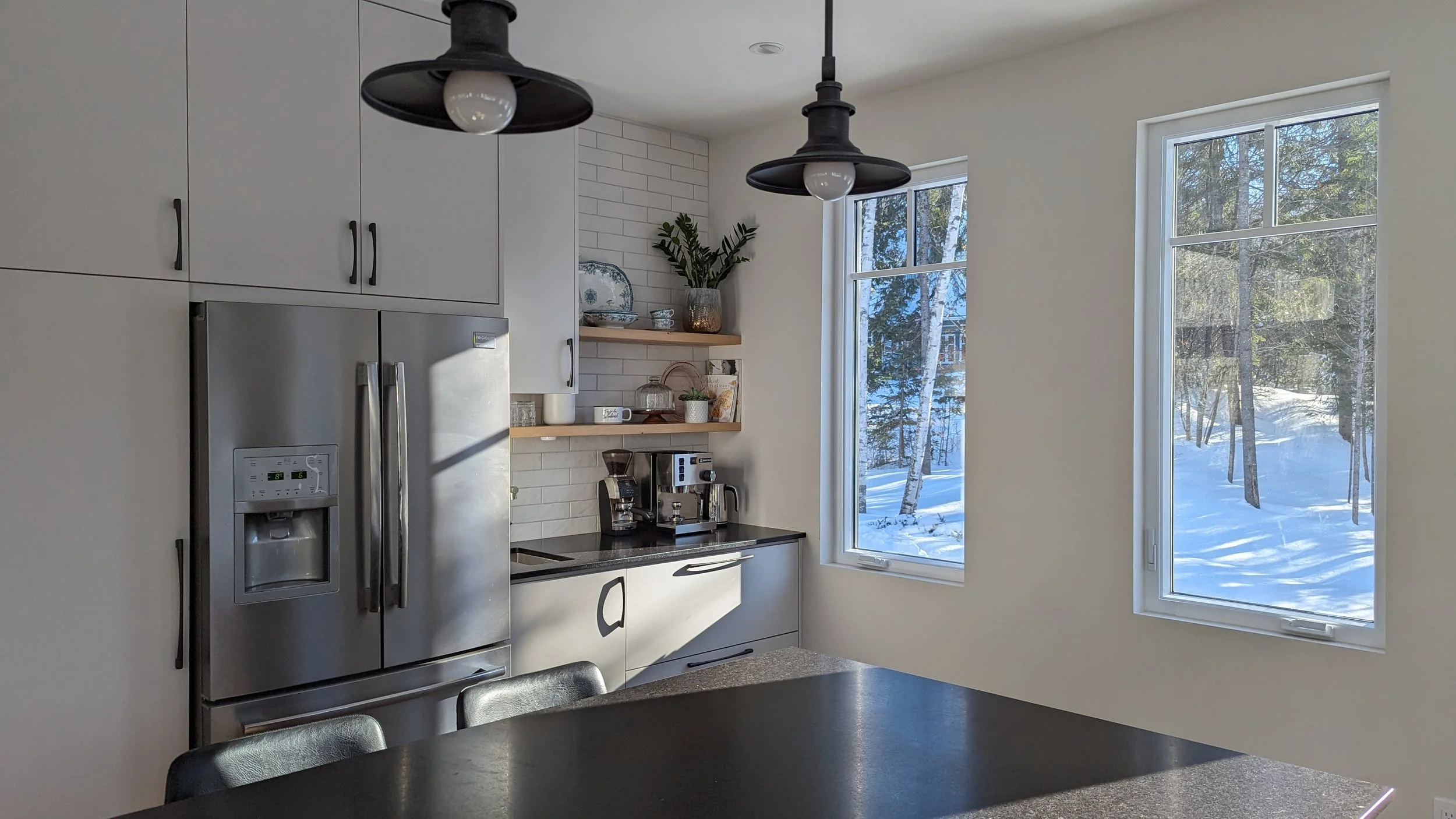 Modern kitchen with white cabinets, black countertop, stainless steel refrigerator, open shelves with dishes and plants, black pendant lights, large windows showing snowy outdoor scene, and black dining table with chairs.