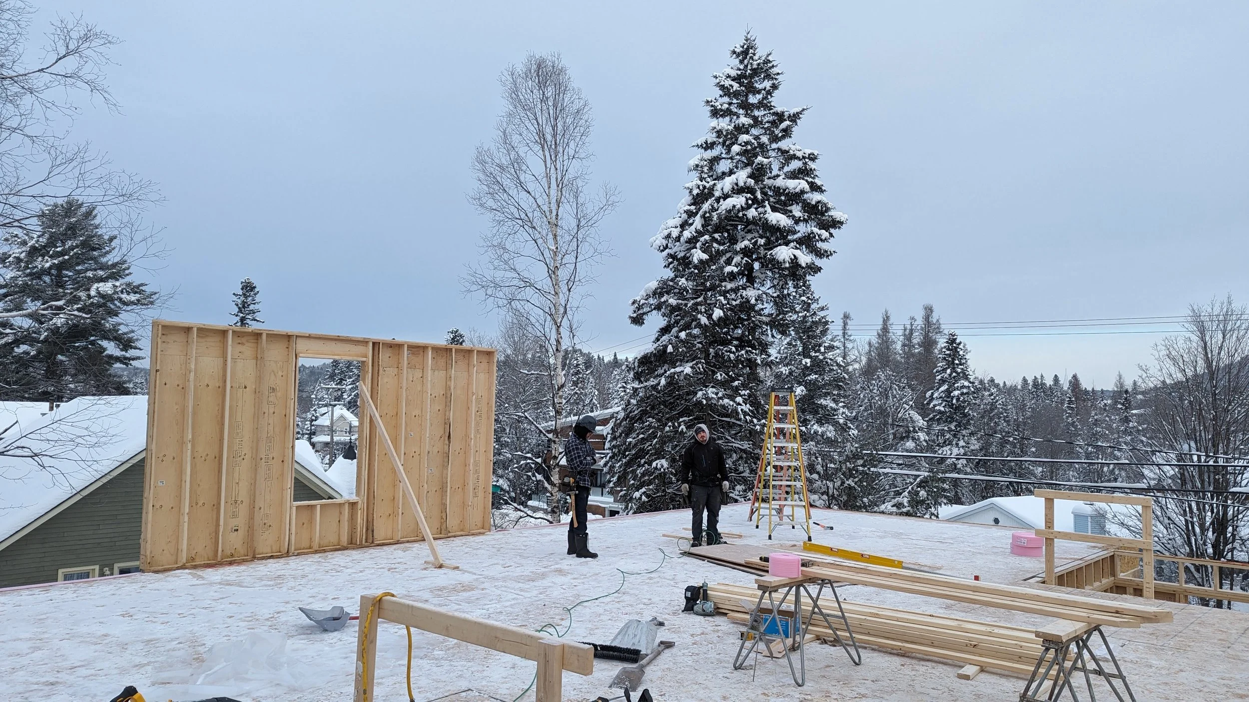 Construction workers building a wooden structure on a snowy rooftop with trees and houses in the background.
