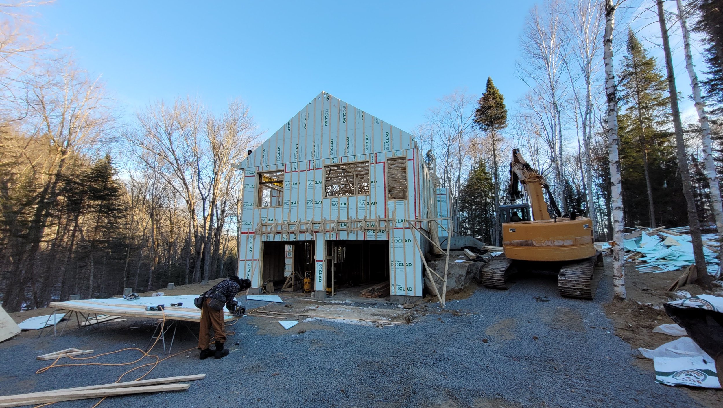 Construction site with a partially built two-story house, surrounded by trees. A worker is measuring wood on the ground, and a yellow excavator is parked nearby.