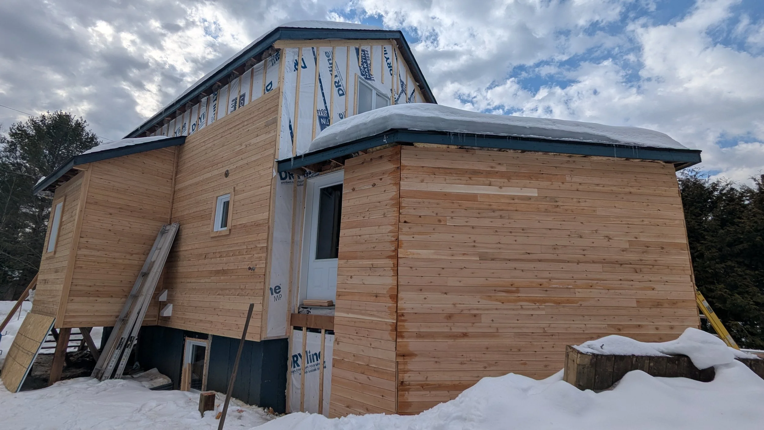 A house under construction with snow on the ground. The exterior walls are partially covered with wooden siding, and some areas of the house are still under insulation. The roof has a layer of snow, and there are windows and a door installed. Construction tools and materials are visible around the house.