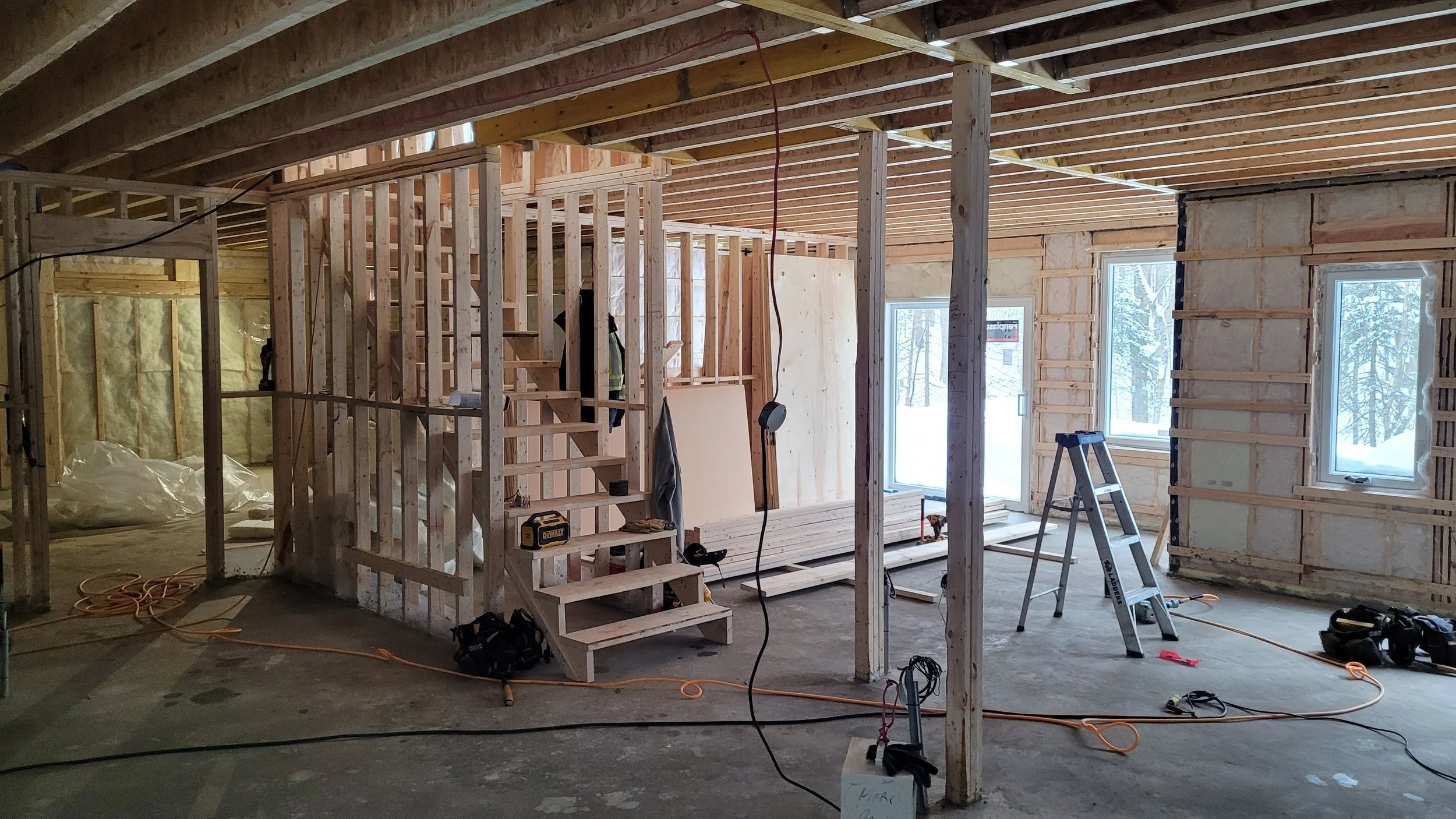 Interior of a house under construction with exposed wooden framing, stairs, and drywall partially installed, with construction tools and ladders on the floor.