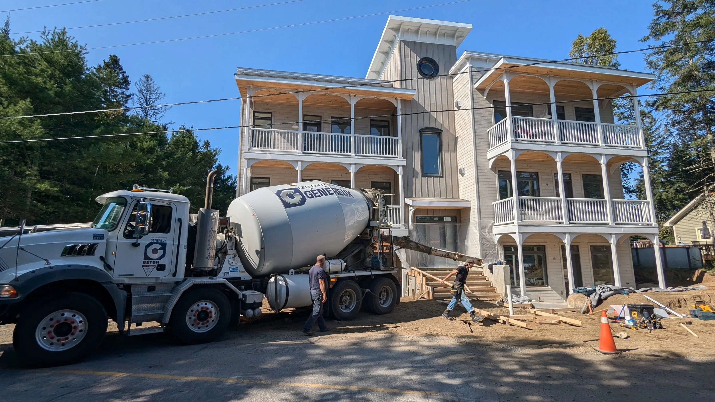 A construction site in front of a three-story house with balconies, where workers are pouring concrete from a cement mixer truck.