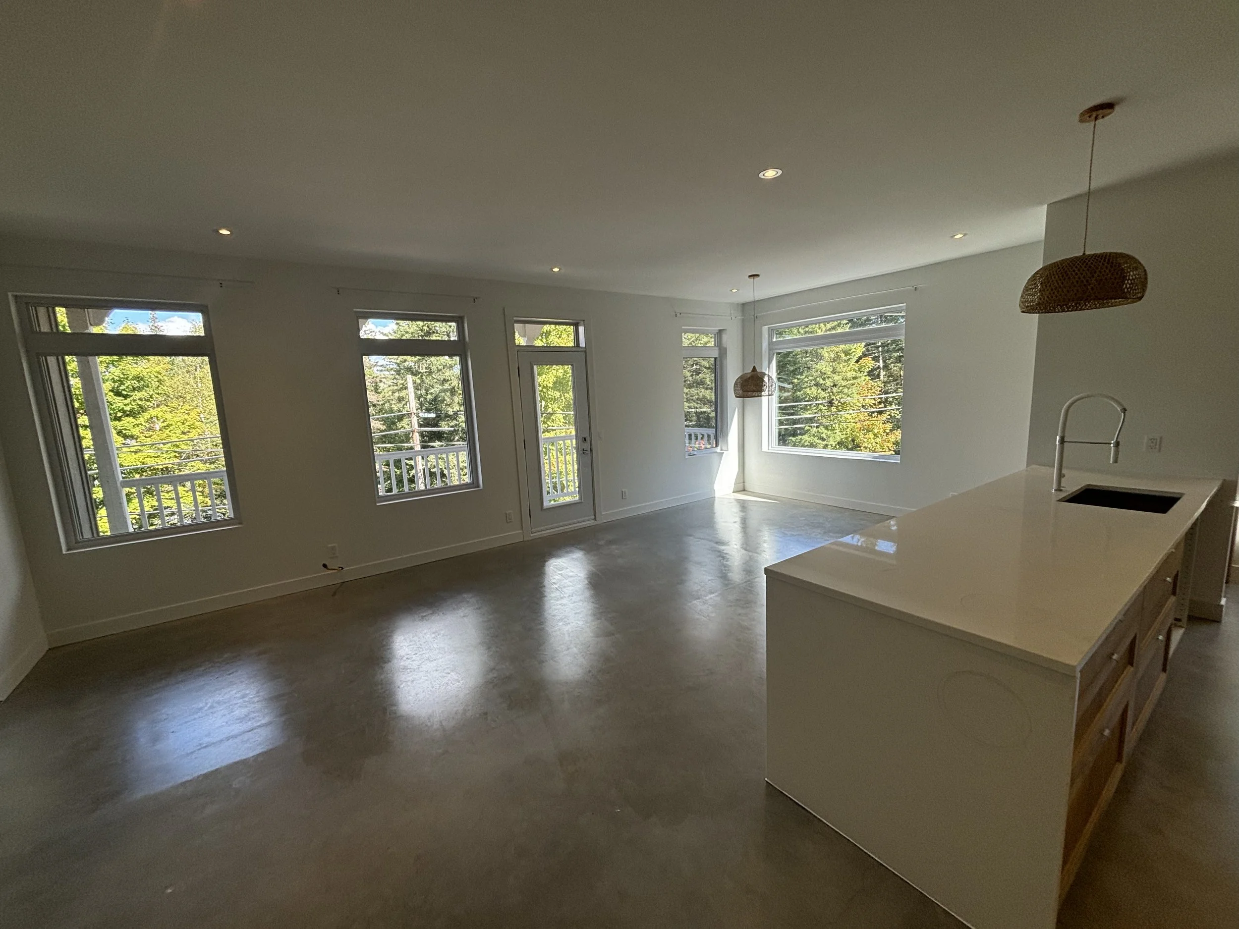 Empty living space with large windows, hardwood floors, and a kitchen island with a sink, pendant lights, and white walls.