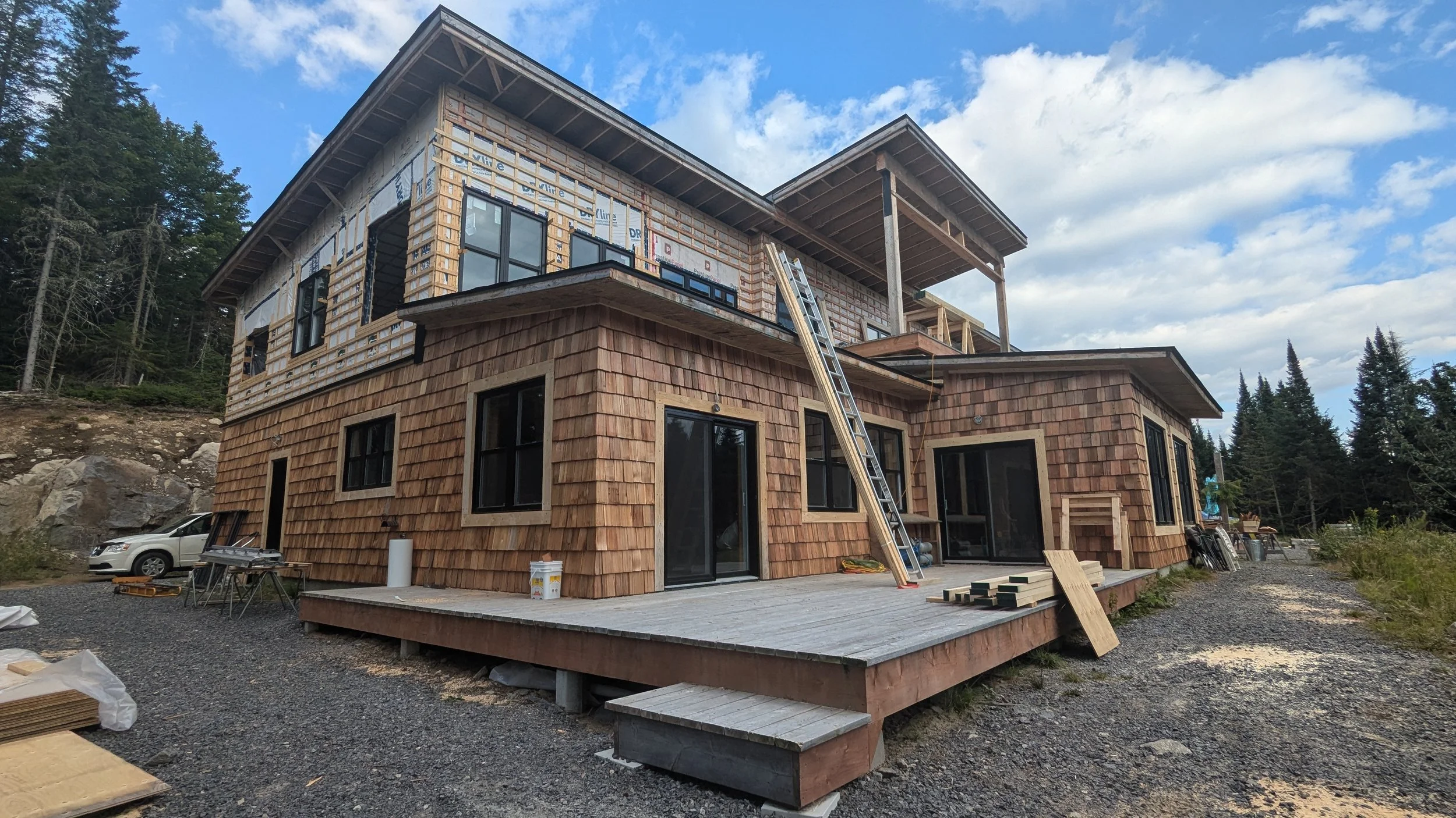 A multi-story house under construction with wooden siding, large windows, and a deck, surrounded by trees and a gravel yard.
