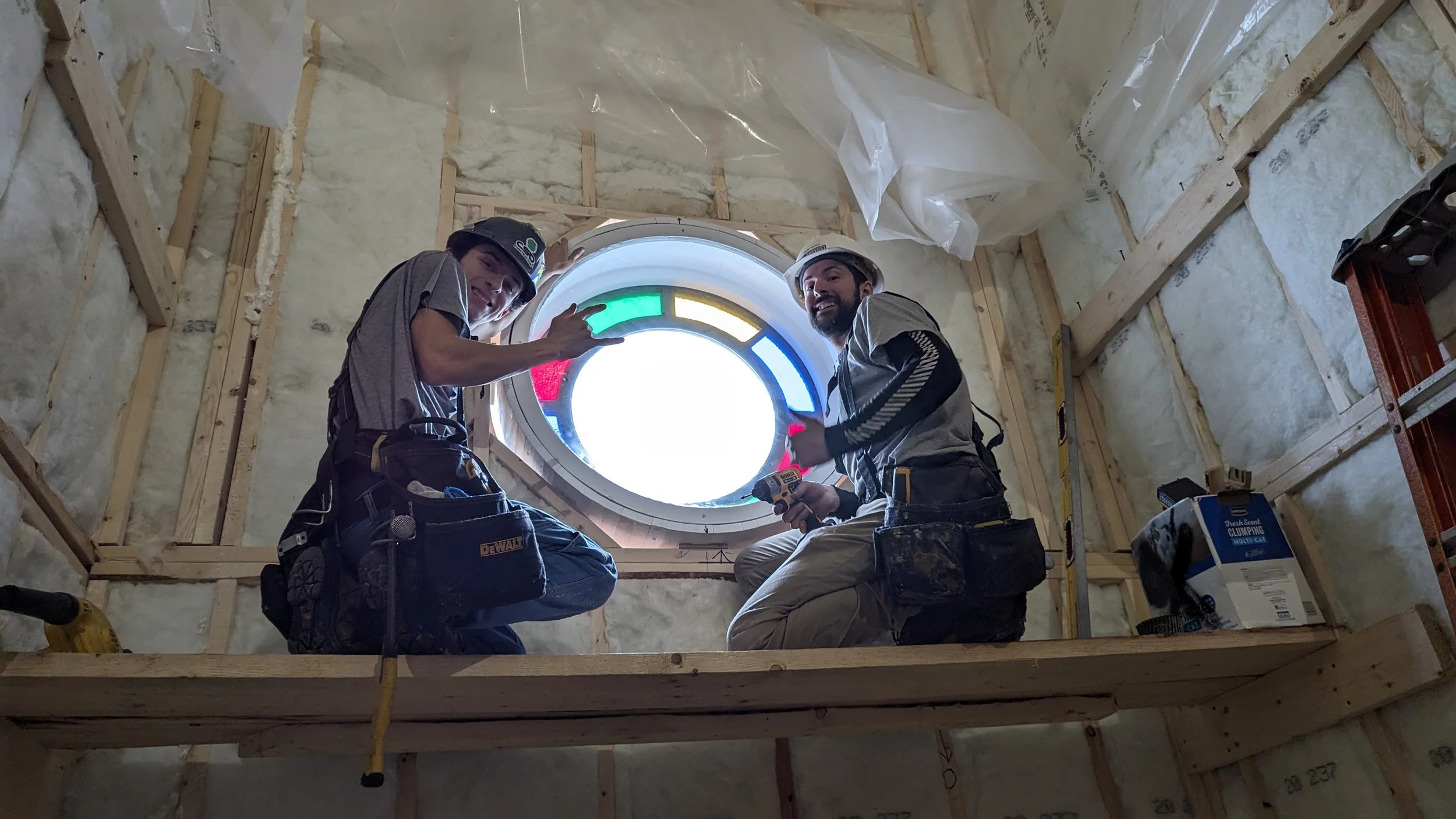 Two construction workers inside a building attic near a circular window, working on installation or maintenance, with tools and construction equipment around them.