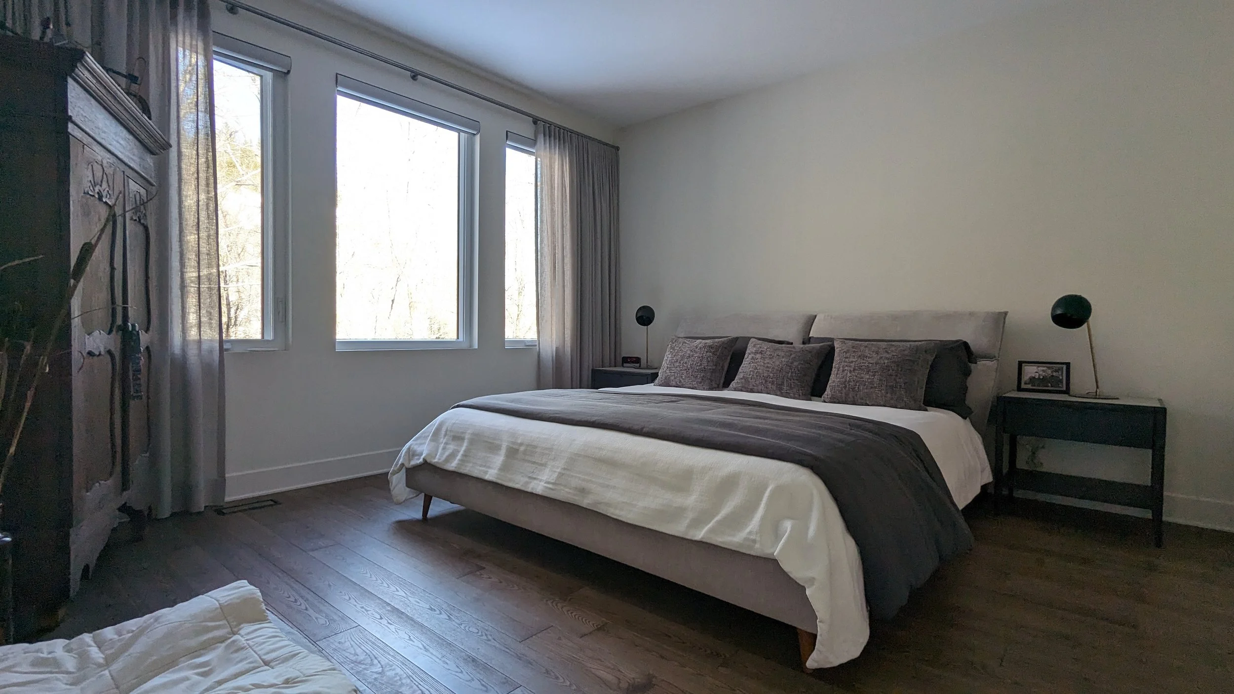 A bedroom with large windows, a bed with dark and light pillows, two black bedside tables with black lamps, a wooden dresser, curtains, and wooden flooring.