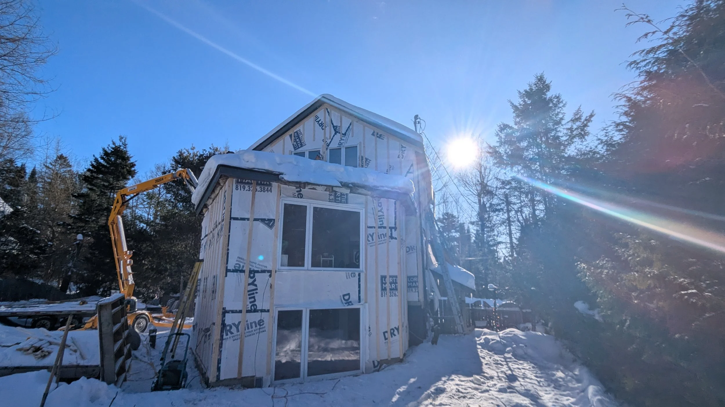 A house under construction with snow on the ground, surrounded by trees, with the sun shining brightly in a clear blue sky.