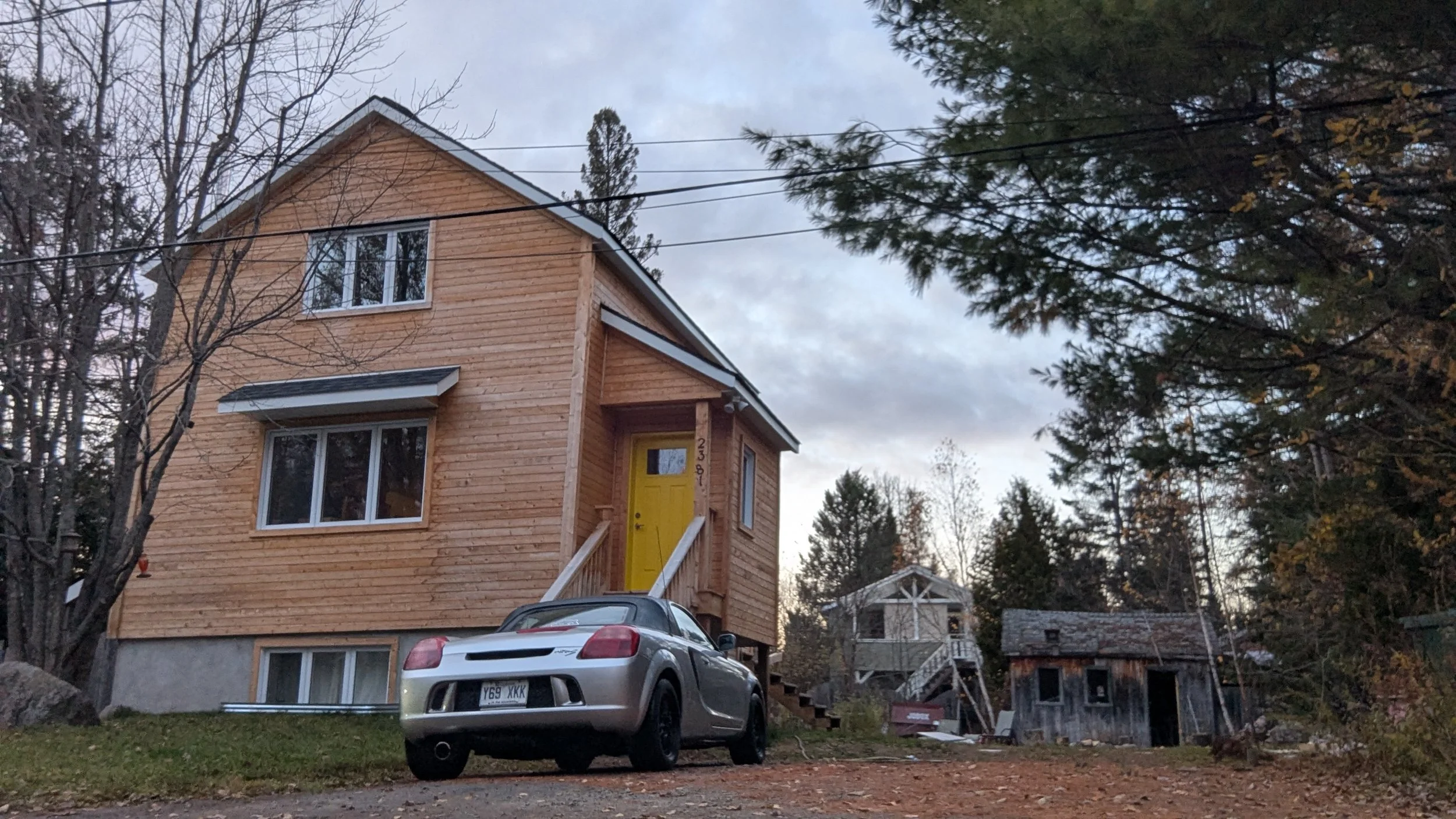 A newly built wooden house with a bright yellow front door, a silver sports car parked in front, and a small weathered shed to the right among trees and cloudy sky.