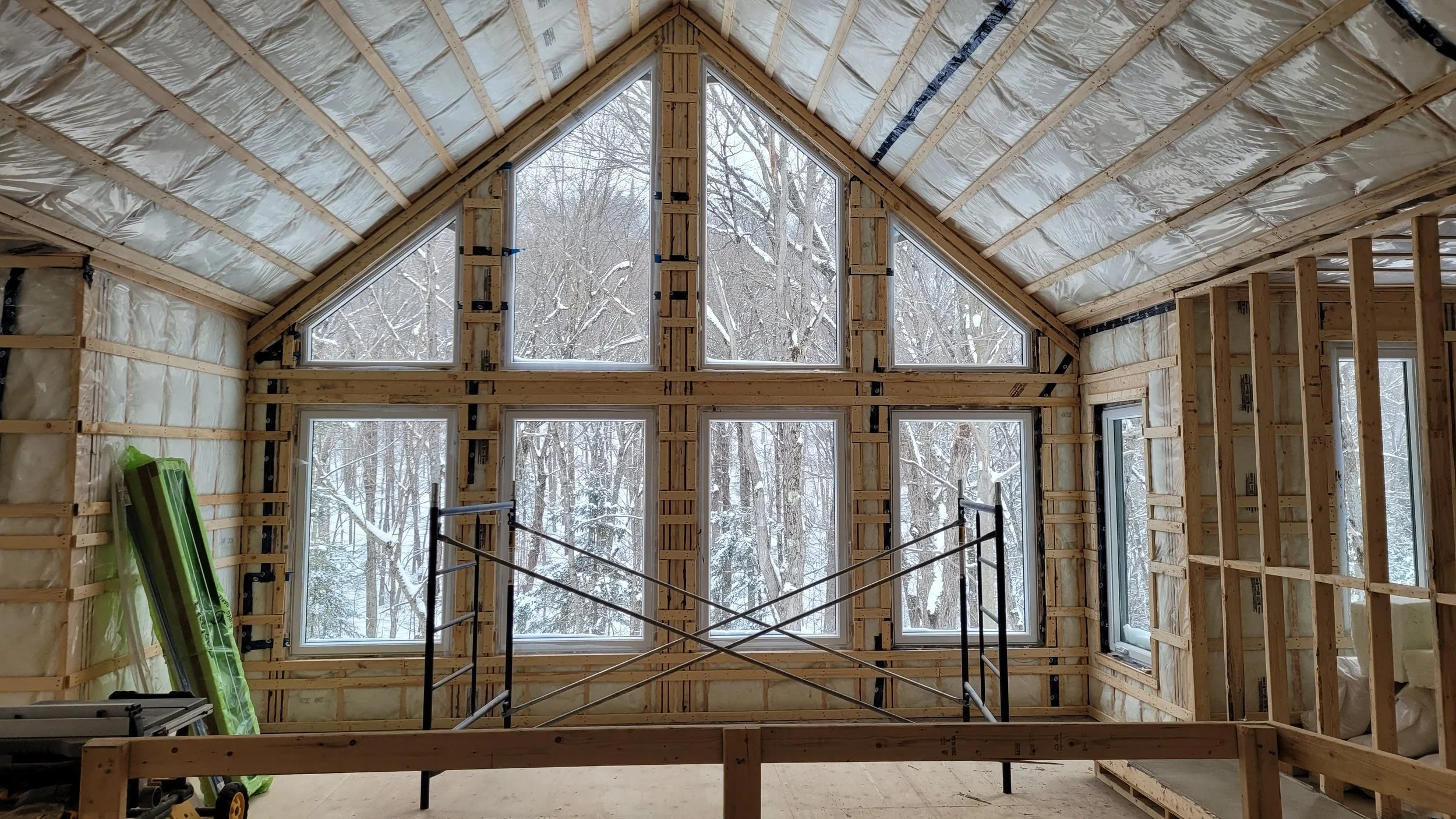Interior view of a house under construction with exposed wooden framing, large multi-pane windows, and snow-covered trees visible outside.
