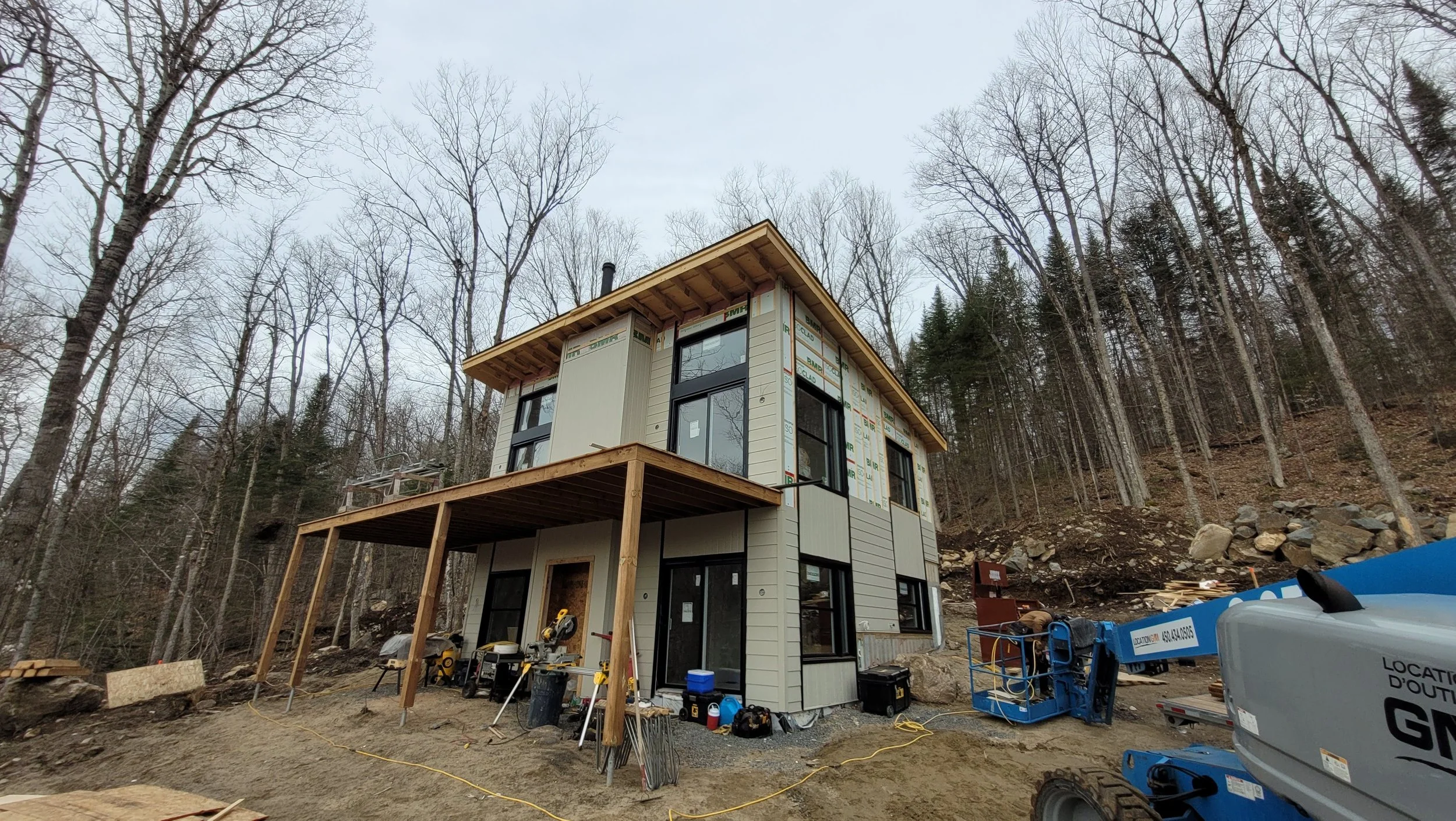 A multi-story house under construction in a wooded area, with a wooden deck porch, construction tools, and equipment around.
