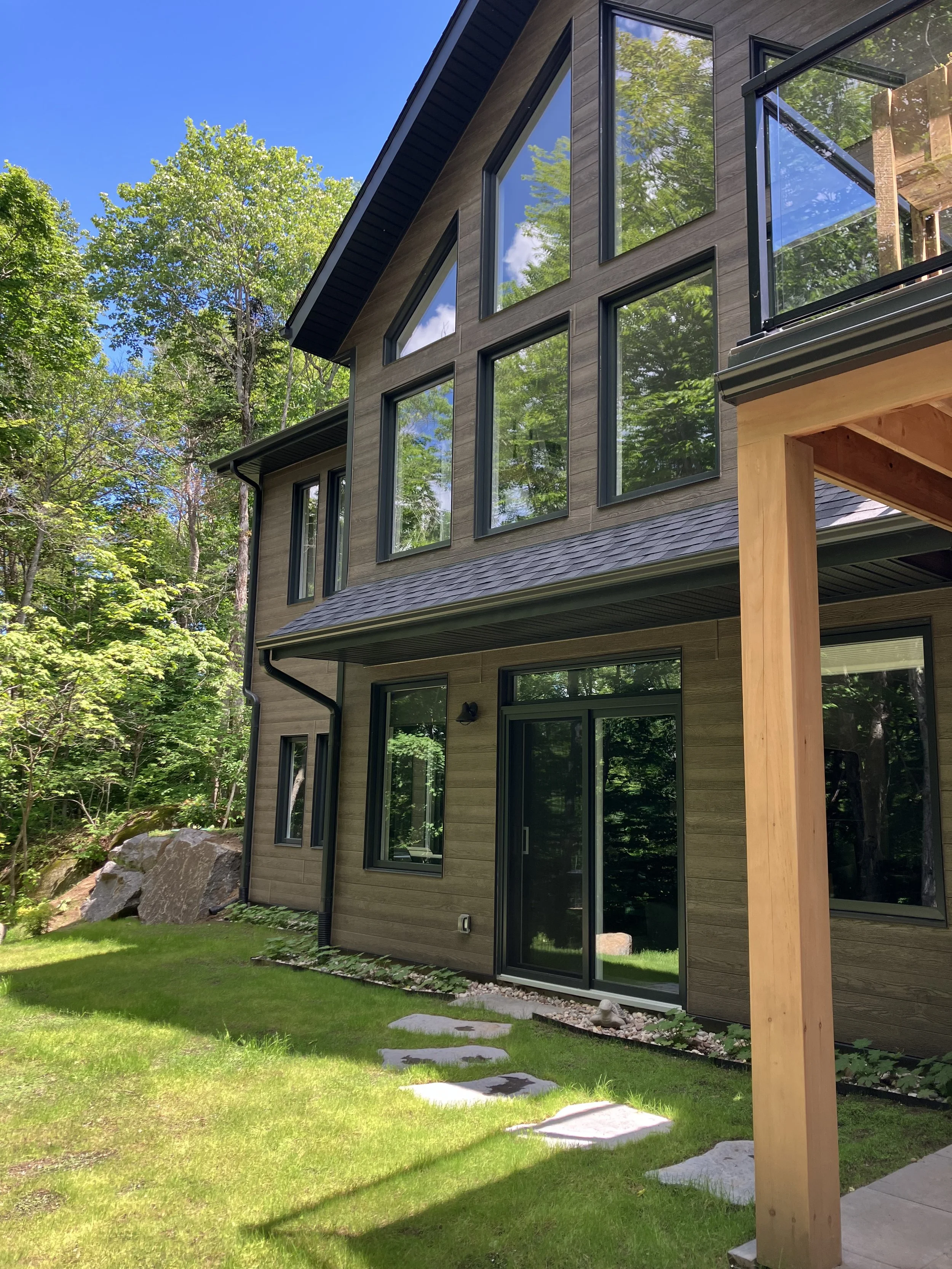 A modern, multi-story house with large windows, wooden exterior, and a glass door, surrounded by green grass and trees under a bright blue sky.
